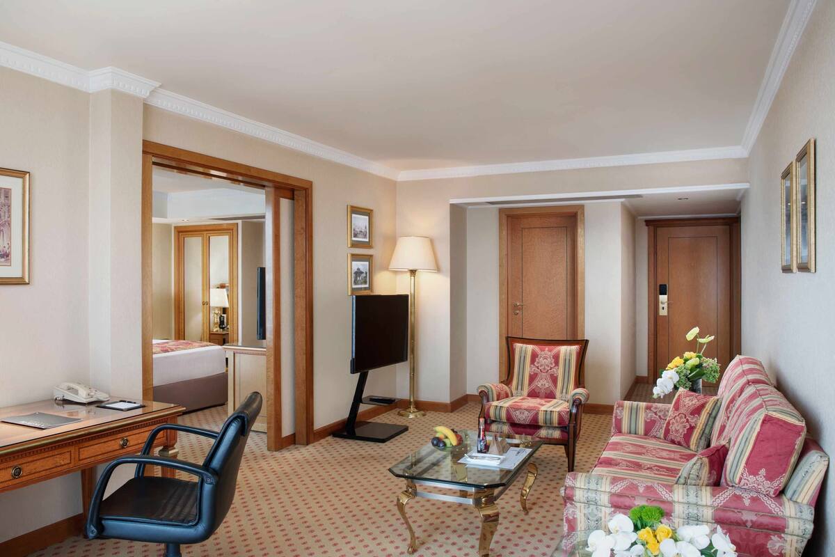 A hotel room with a desk, chair, sofa, TV, and a mirror.