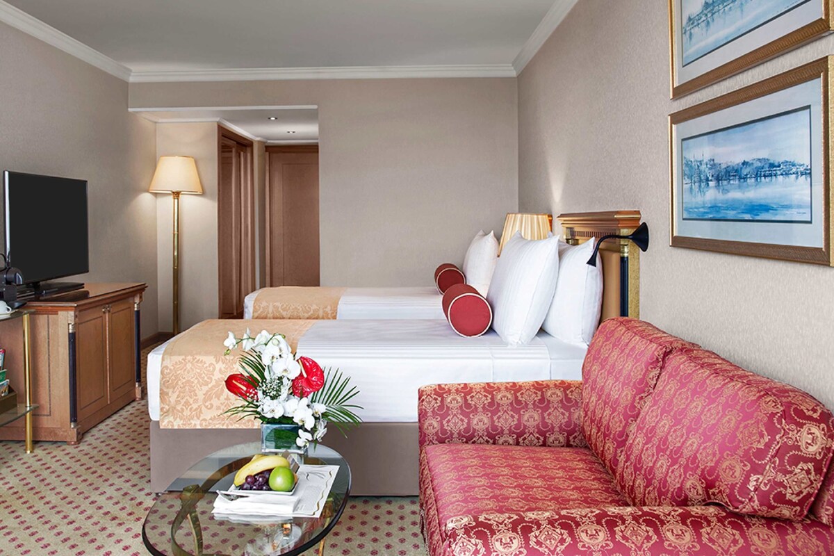 A hotel room with a bed, a sofa, a TV, a desk with a lamp, and a coffee table with a book and flowers.