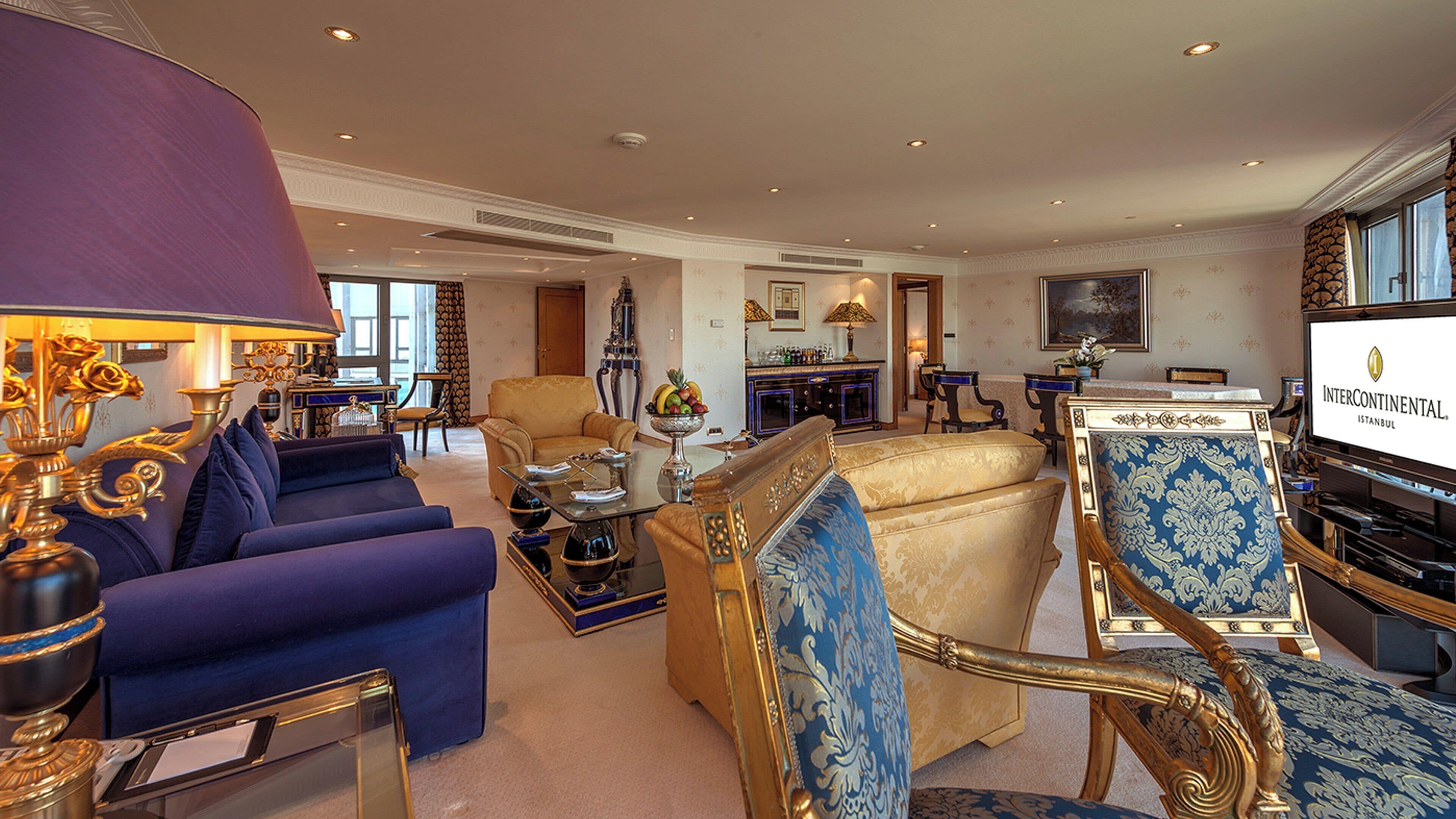 A spacious living area with a large sofa, a coffee table, and a television. There is a dining area with a table and chairs, and a kitchen in the background.