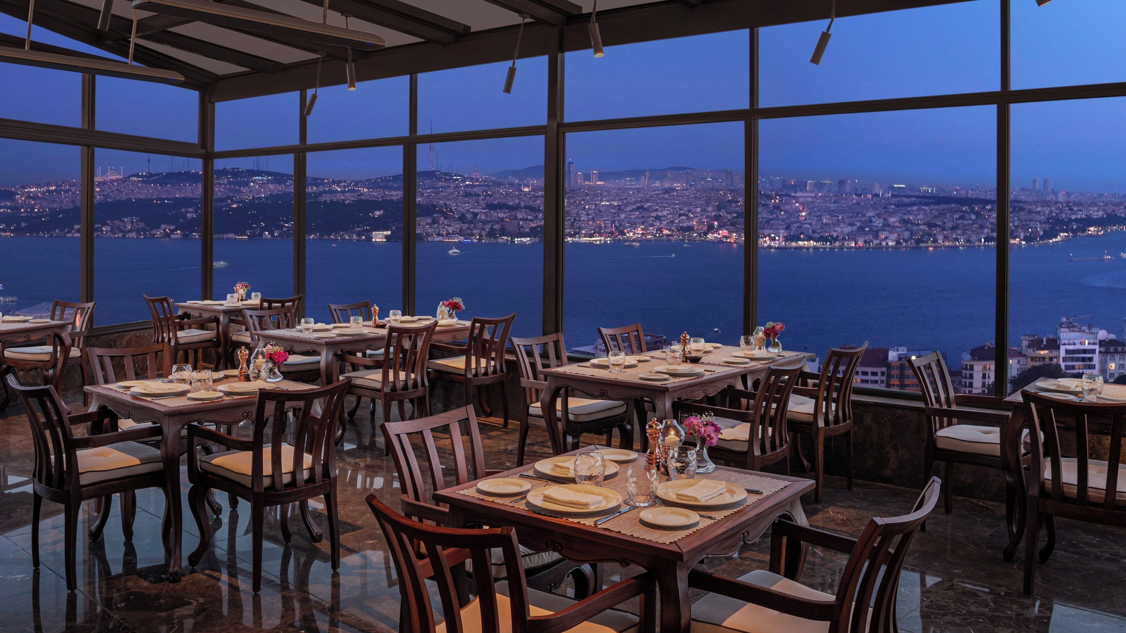 Un restaurante con varias mesas dispuestas para comer, amplias ventanas que ofrecen vistas de la ciudad y una decoración de estilo moderno.
