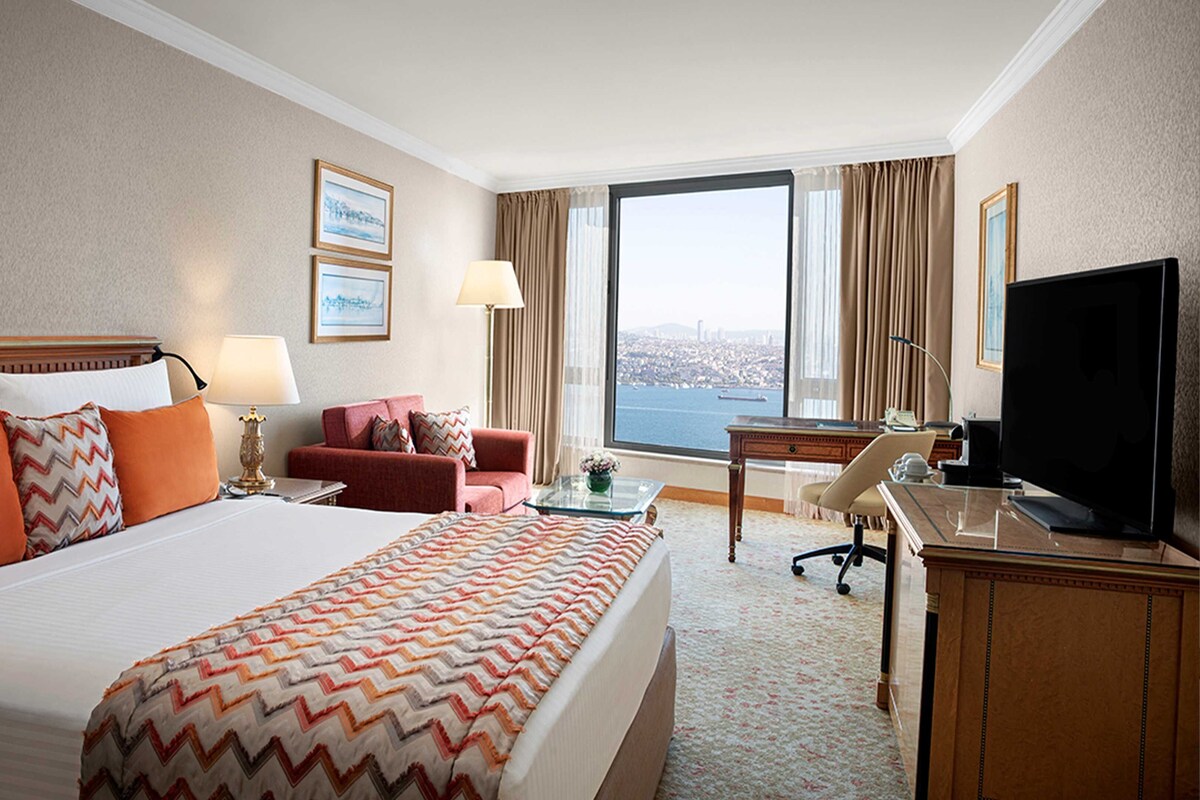A hotel room with a large bed, a desk, a chair, a television, and a view of the city through the window.
