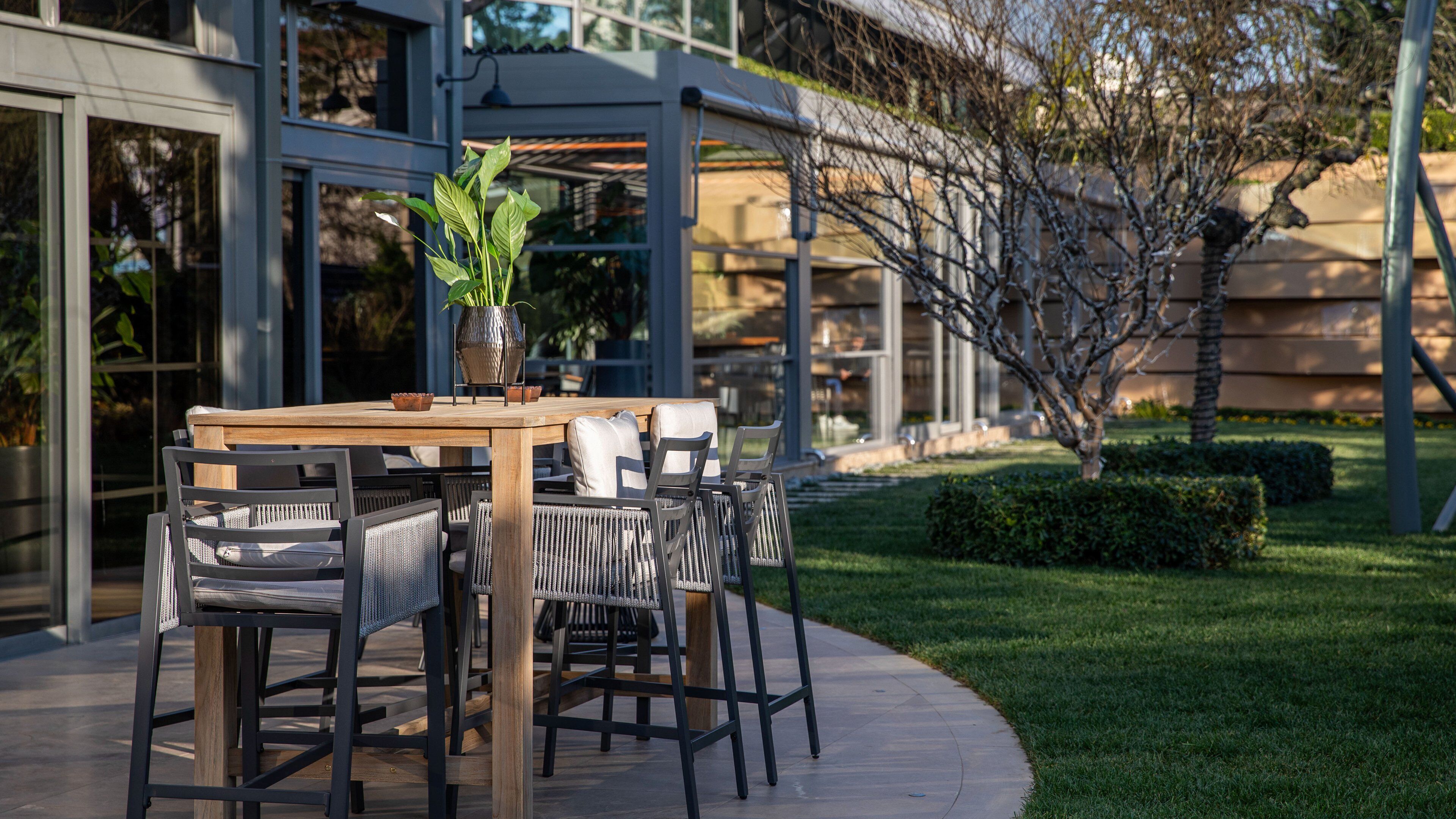 An outdoor dining area with a wooden table and chairs, a potted plant, and a modern building in the background.