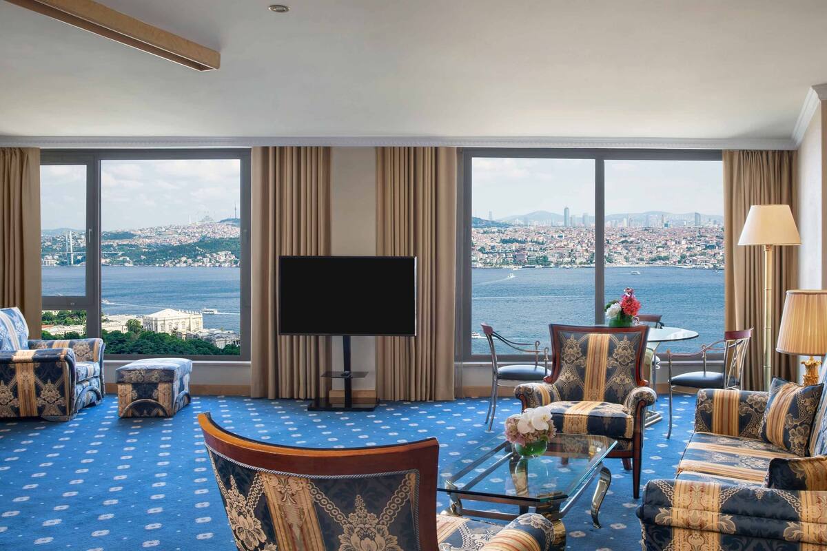 A spacious hotel room with a large window offering a cityscape view, a flat-screen TV, and a dining area with a table and chairs.