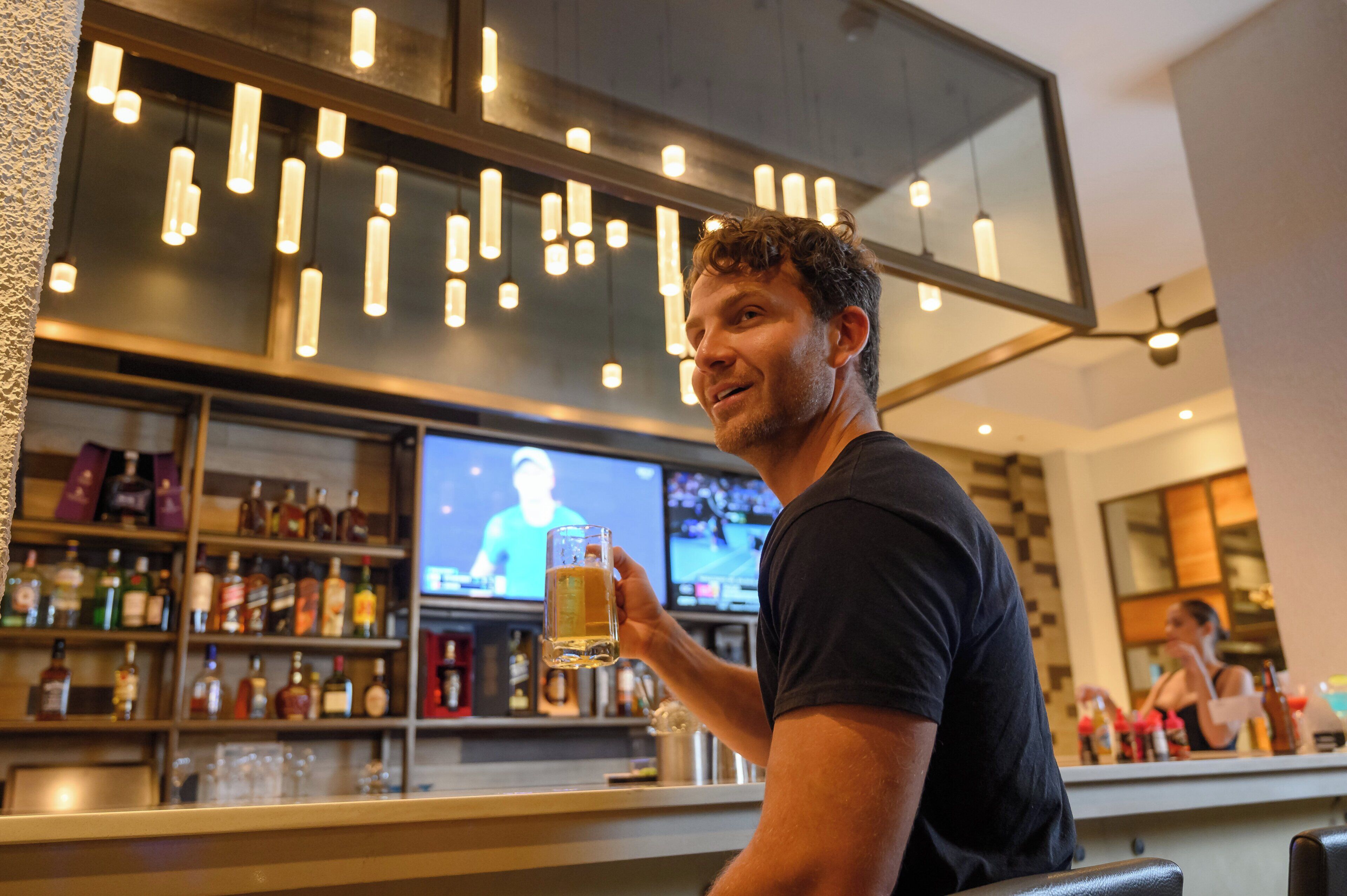 A man holding a beer glass in a bar with shelves of liquor bottles and a television screen in the background.