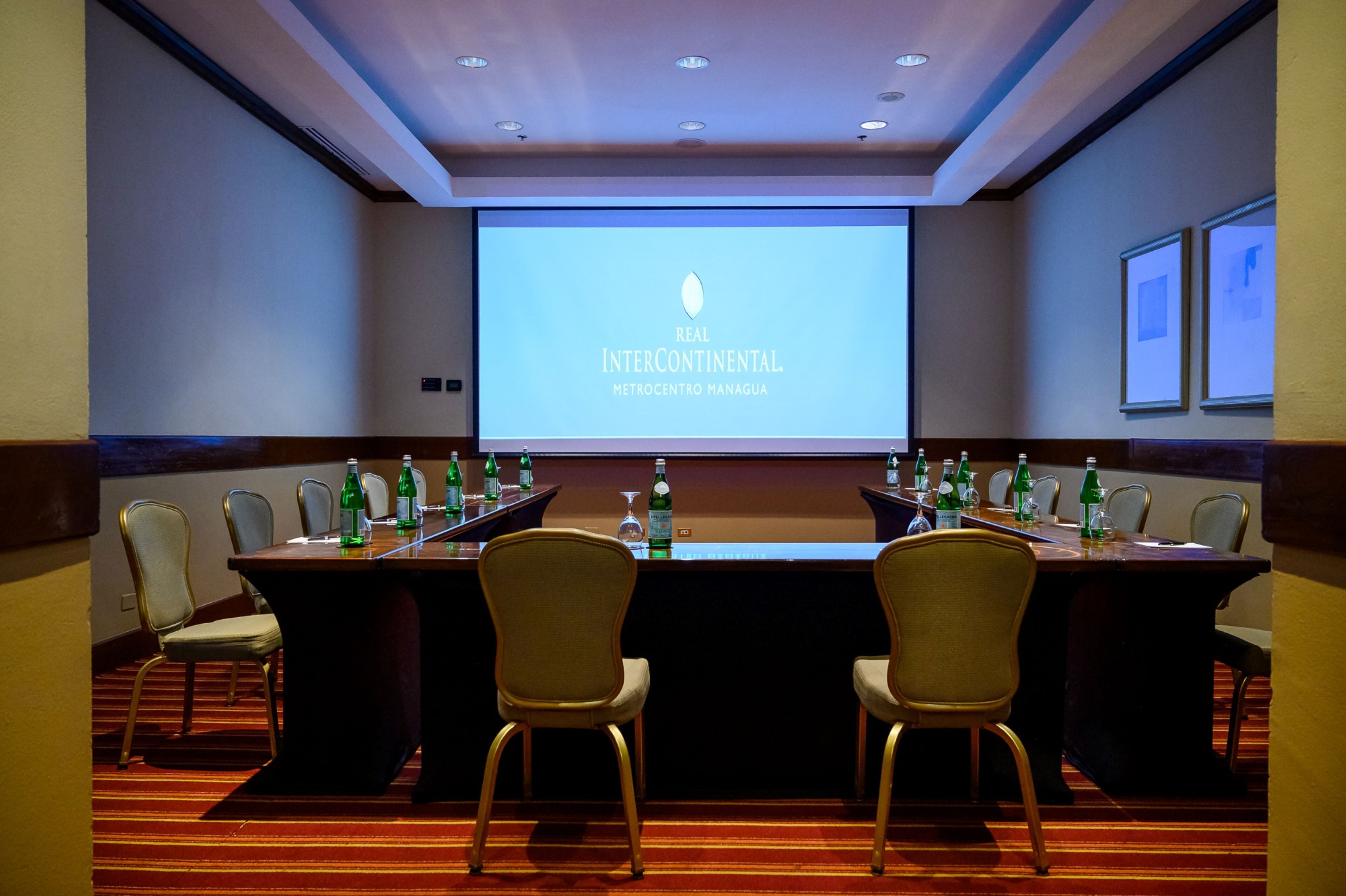 A conference room with a large screen displaying 'INTERCONTINENTAL METROCENTRO MANAGUA' and a long table with chairs and water bottles.