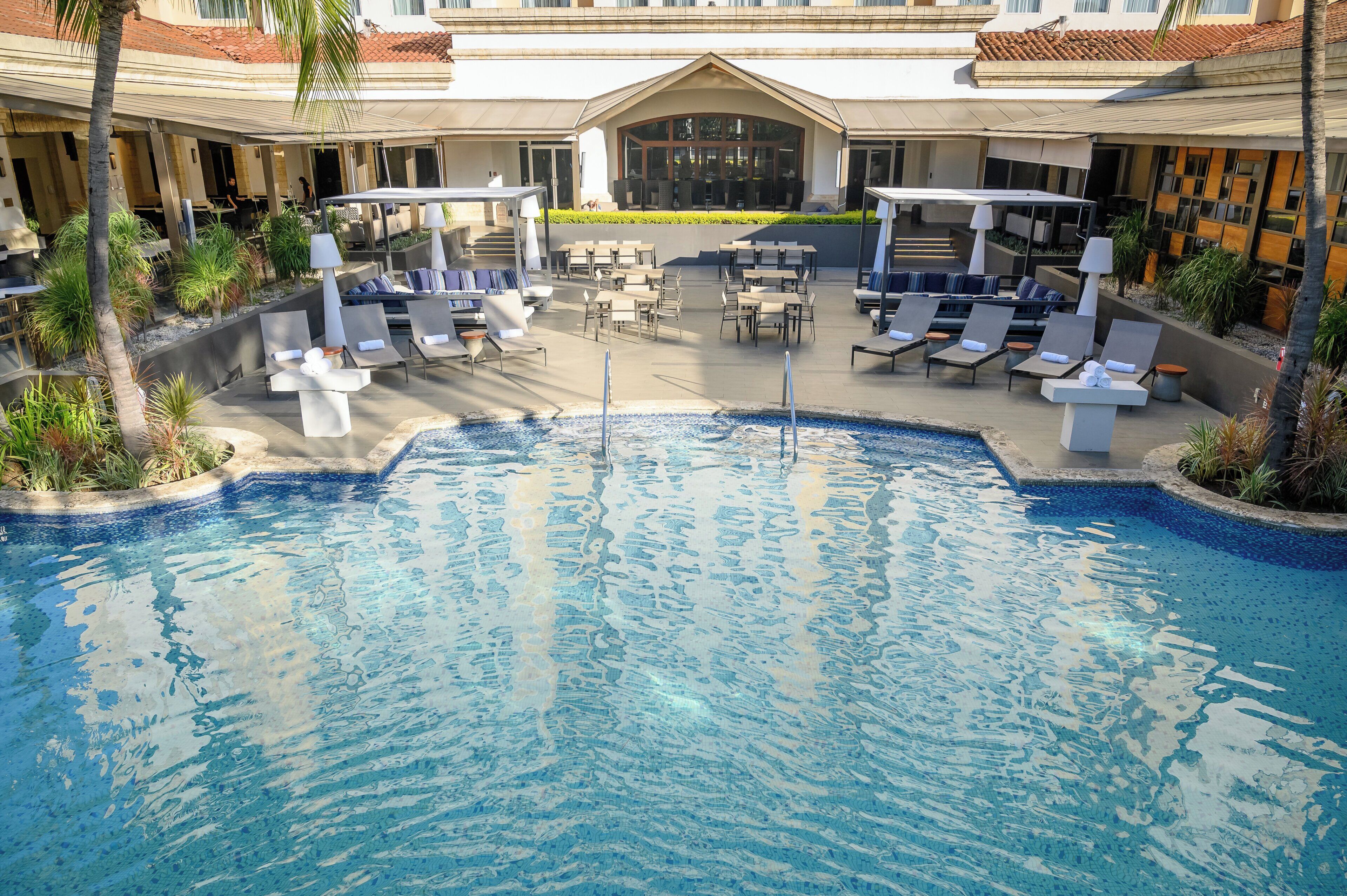 A pool area with lounge chairs and tables, surrounded by a building with a tiled roof and a covered seating area.