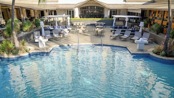 A pool area with lounge chairs and tables, surrounded by a building with a tiled roof and a covered seating area.