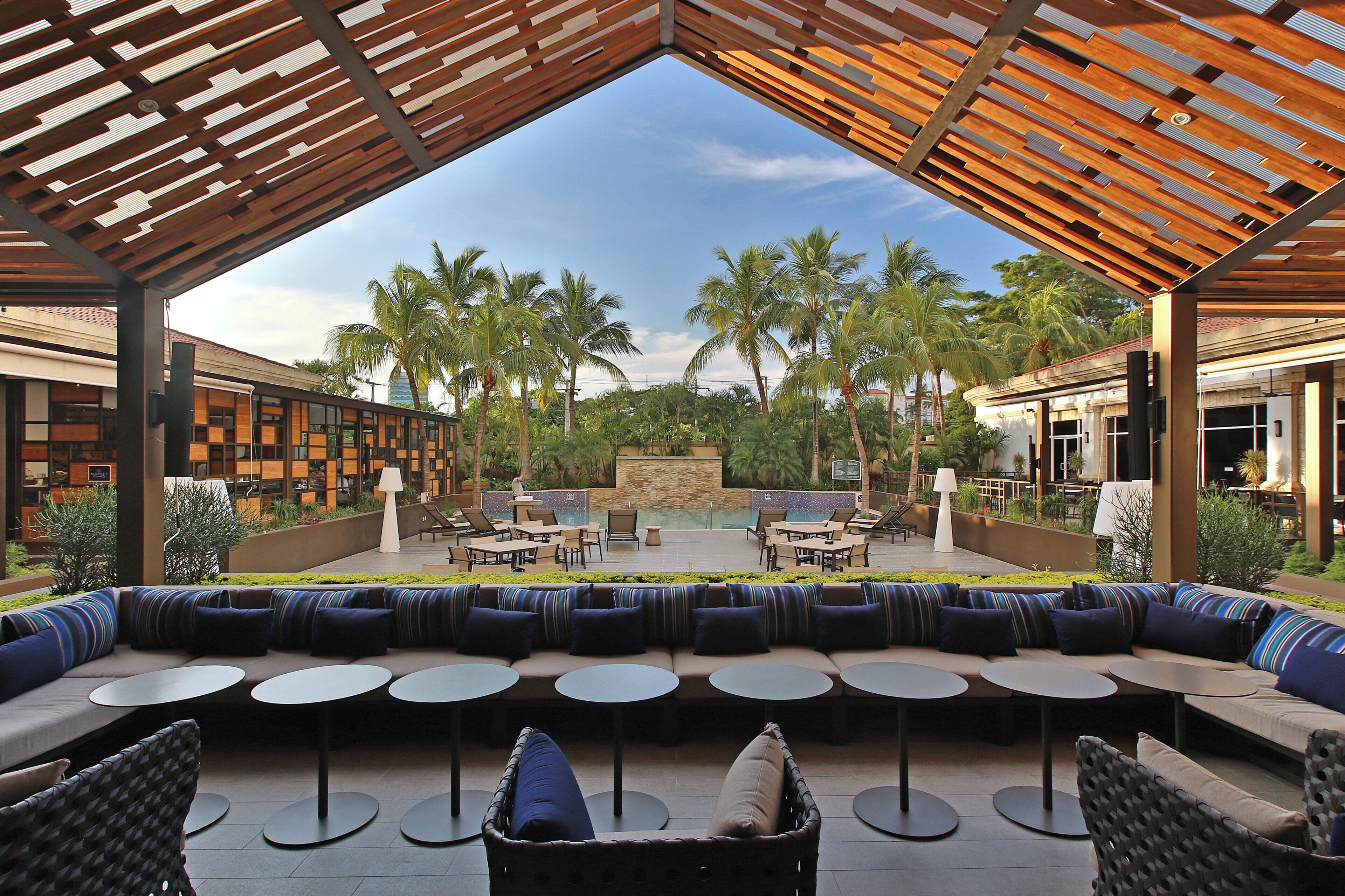 An outdoor seating area with round tables and sofas, a pool, and palm trees.