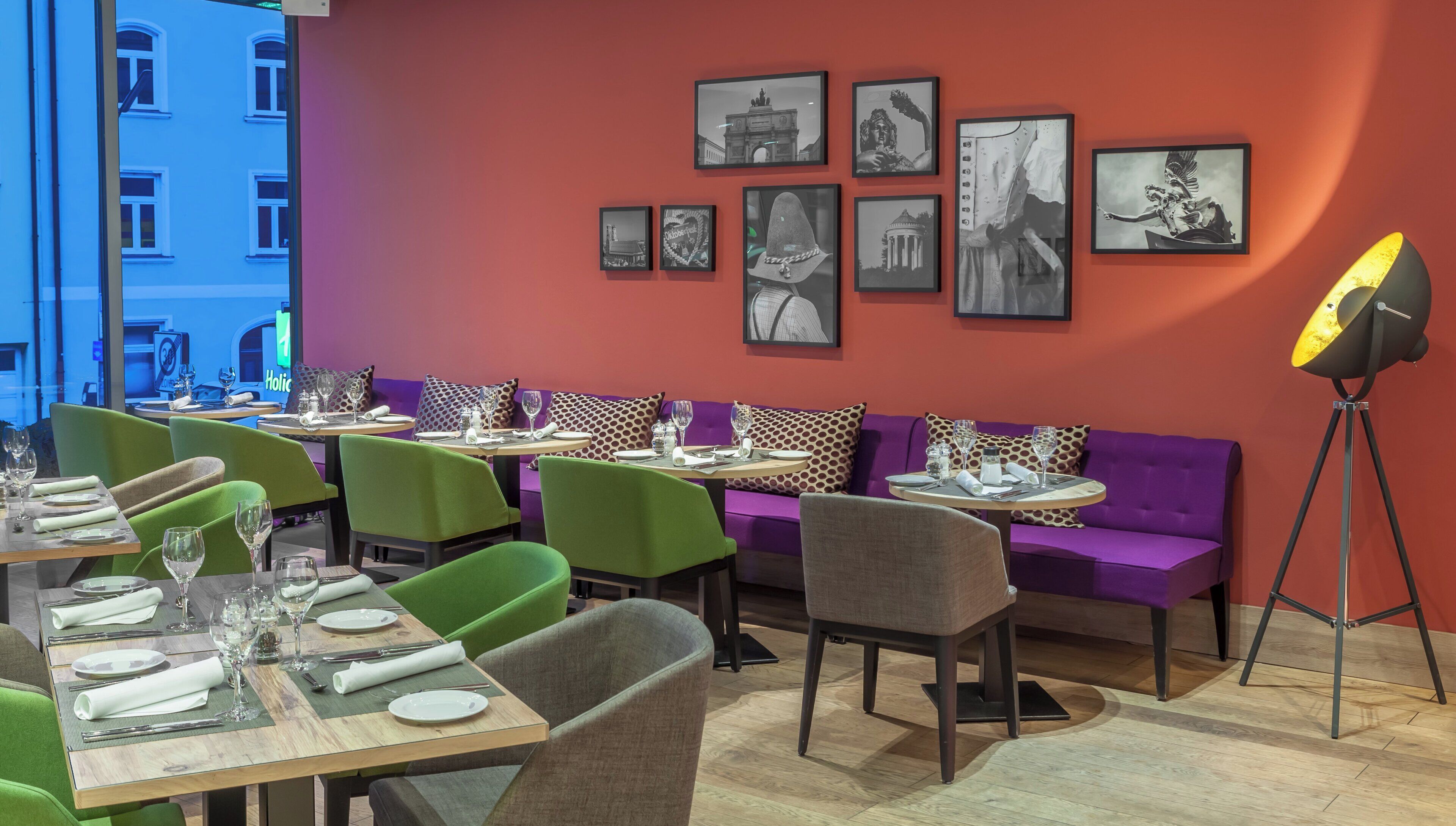 Un restaurante con sillas verdes y sofás morados, mesas dispuestas con platos y vasos, y fotografías en blanco y negro enmarcadas en la pared.