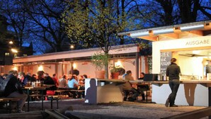 Un área de comedor al aire libre con mesas y sillas, un letrero que dice "AUSGABE" y una persona de pie en el mostrador.