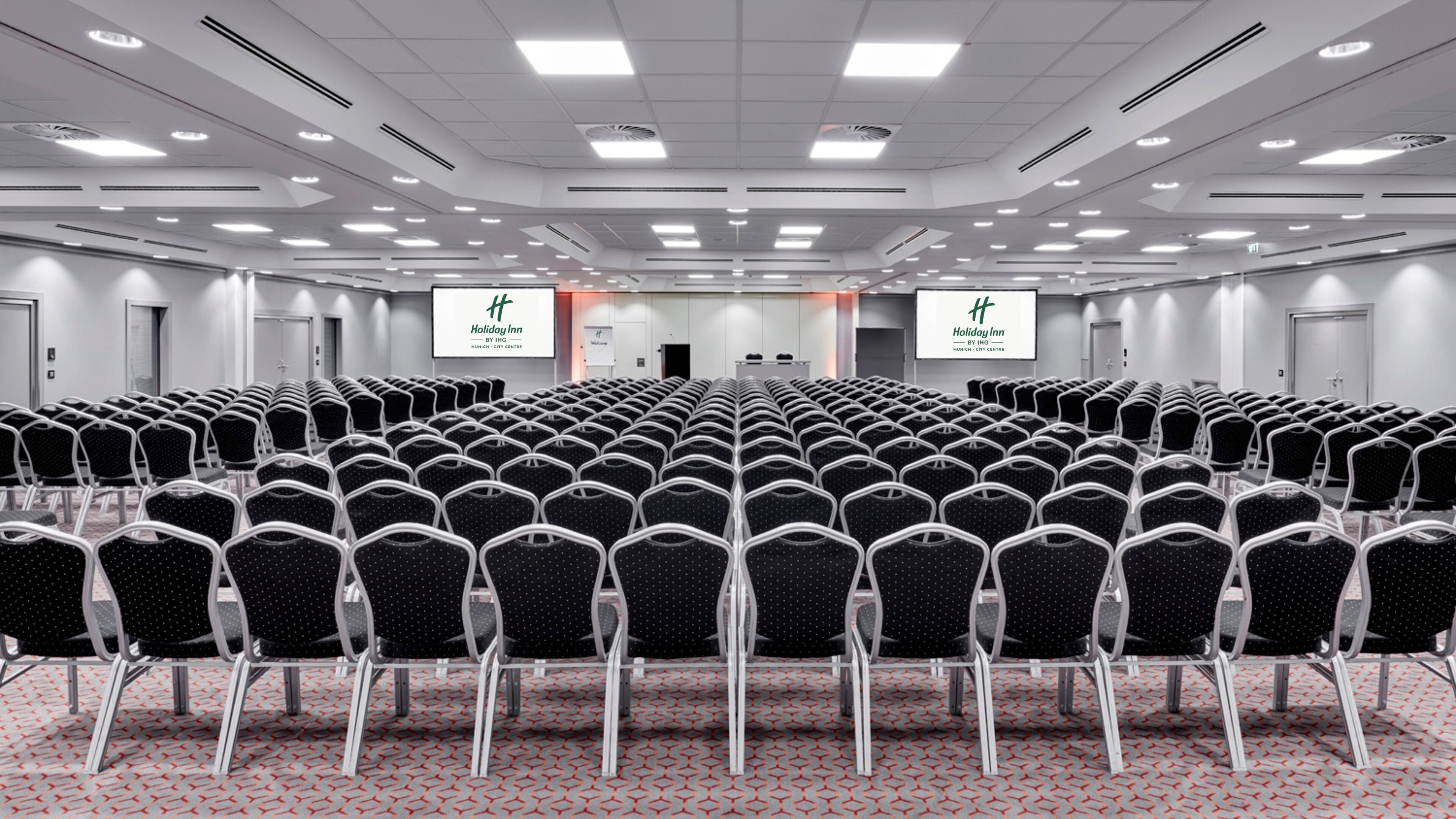 A conference room with rows of chairs, a stage, and two screens displaying the Holiday Inn Munich City Centre logo.