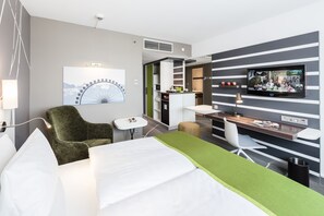 A modern hotel room with a bed, a desk, a TV, a chair, and a small table. - Holiday Inn Munich - City Centre by IHG (Munich)