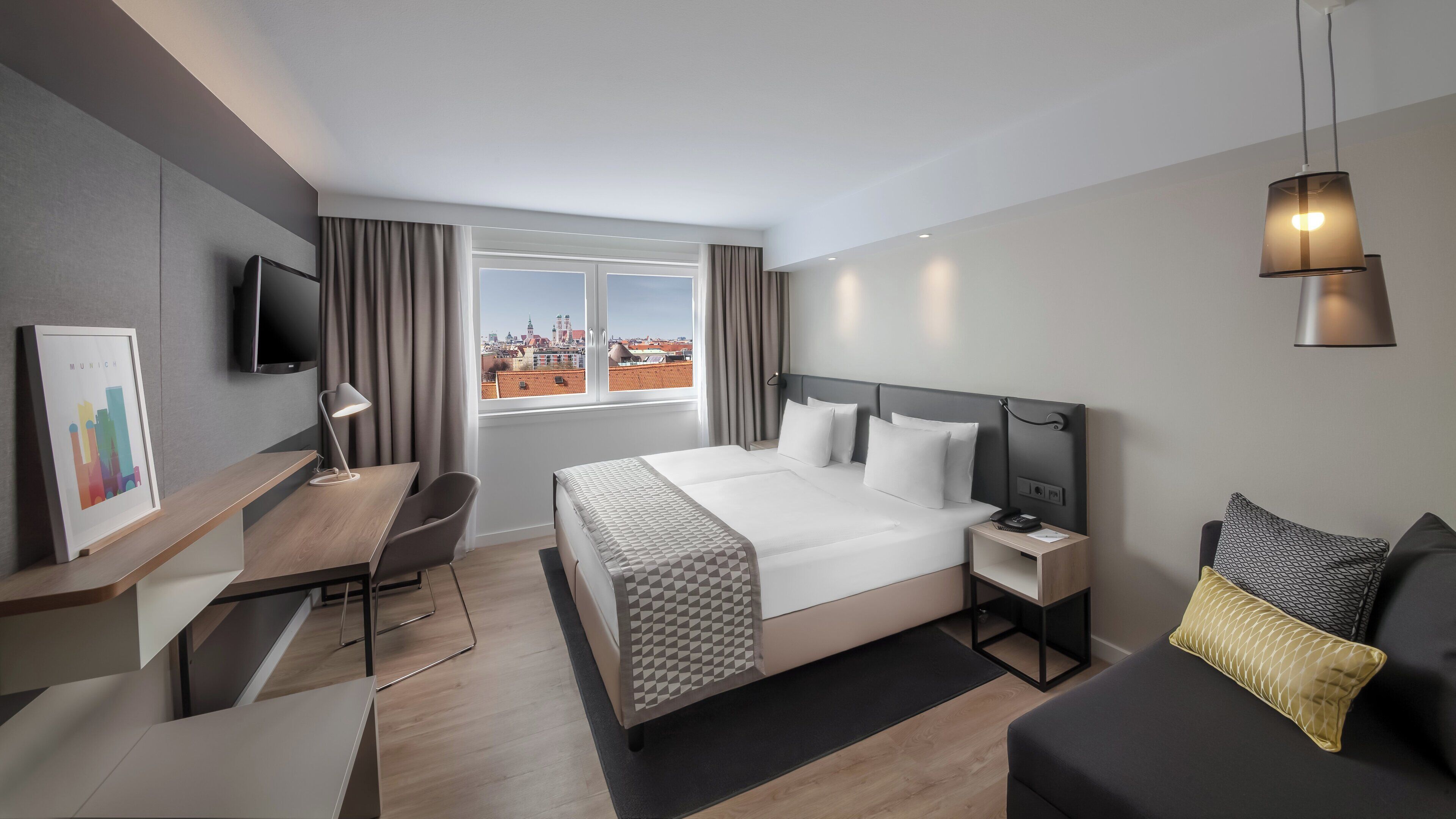 A hotel room with a bed, bedside table, desk, chair, and a view of the city.