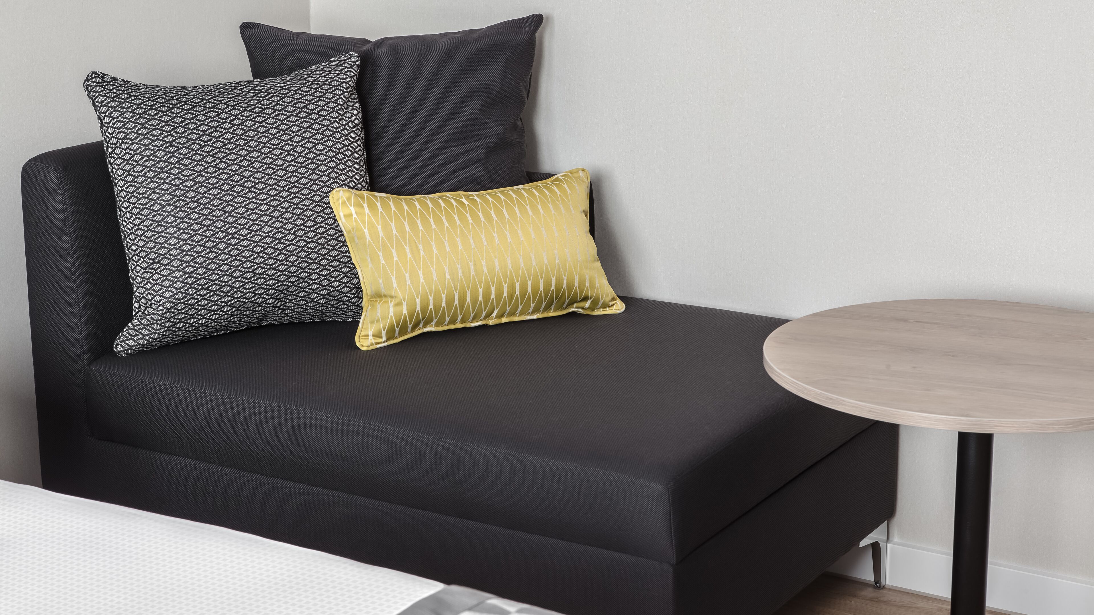 Un sofá moderno con tela oscura, acompañado por dos almohadones estampados, uno con diseño geométrico y el otro con un patrón de cruces. Una pequeña mesa redonda de madera está ubicada a la derecha del sofá.