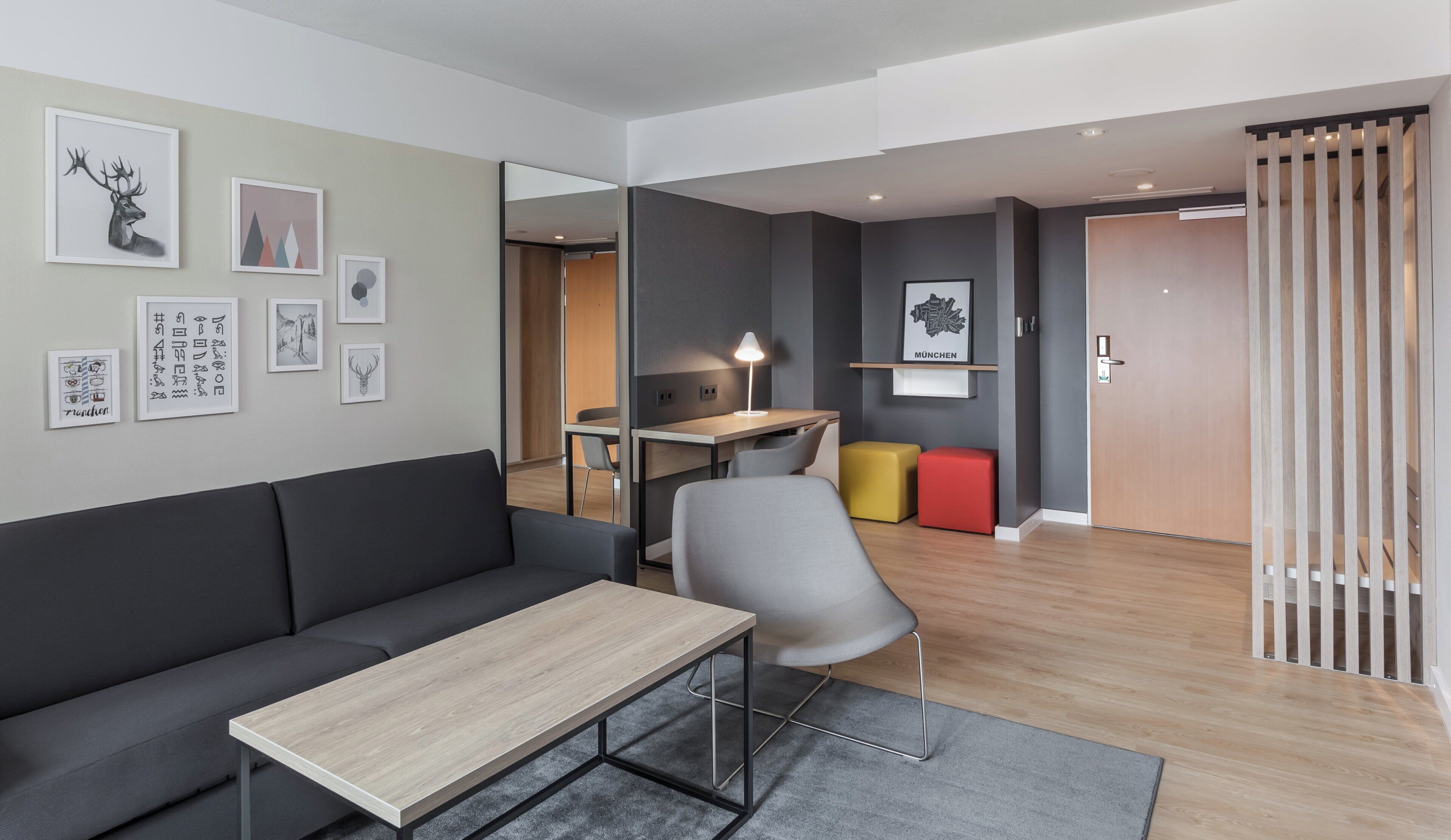 Una sala moderna con un sofá, una mesa de centro y un escritorio con lámpara. Hay cuadros enmarcados en la pared y una puerta con panel de madera.