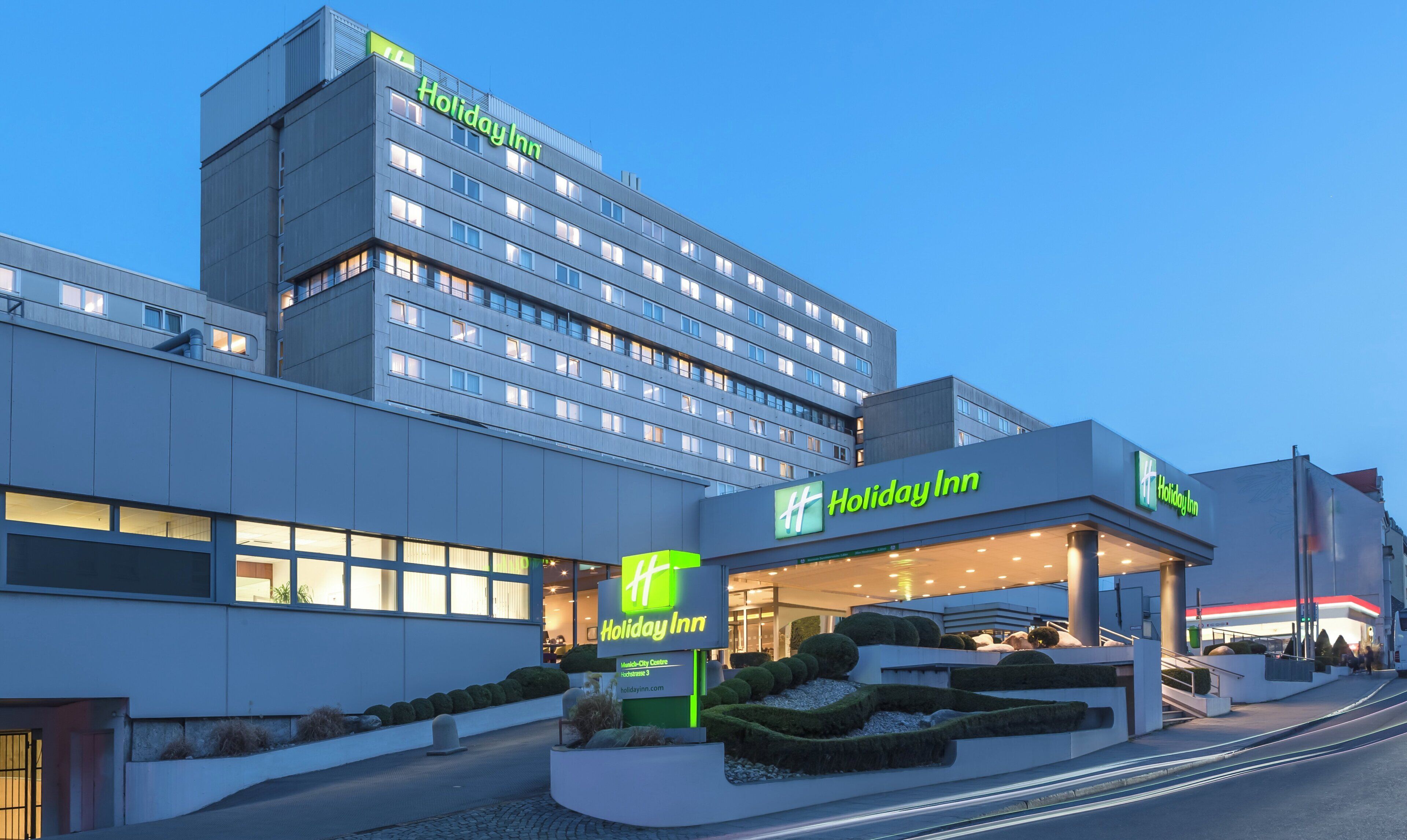 Ein Holiday Inn Hotel mit modernem Design, das ein markantes Schild, einen begrünten Eingangsbereich und ein mehrstöckiges Gebäude mit beleuchteten Fenstern zeigt.