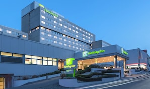 A Holiday Inn hotel with a modern design, featuring a prominent sign, a landscaped entrance, and a multi-story building with illuminated windows. - Holiday Inn Munich - City Centre by IHG (Munich)