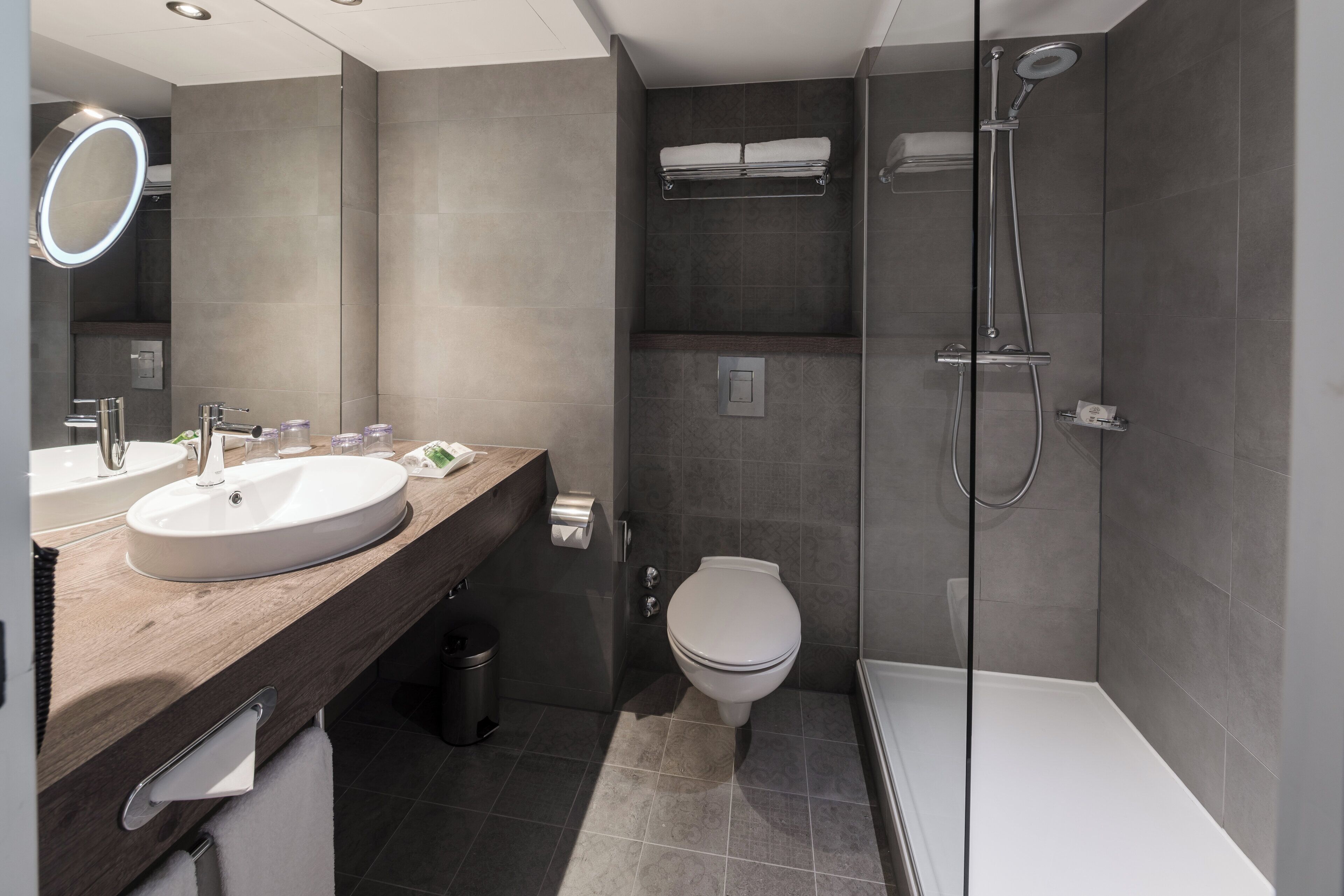 Un baño moderno con tocador de madera, dos lavamanos, inodoro y cabina de ducha con mampara de vidrio.