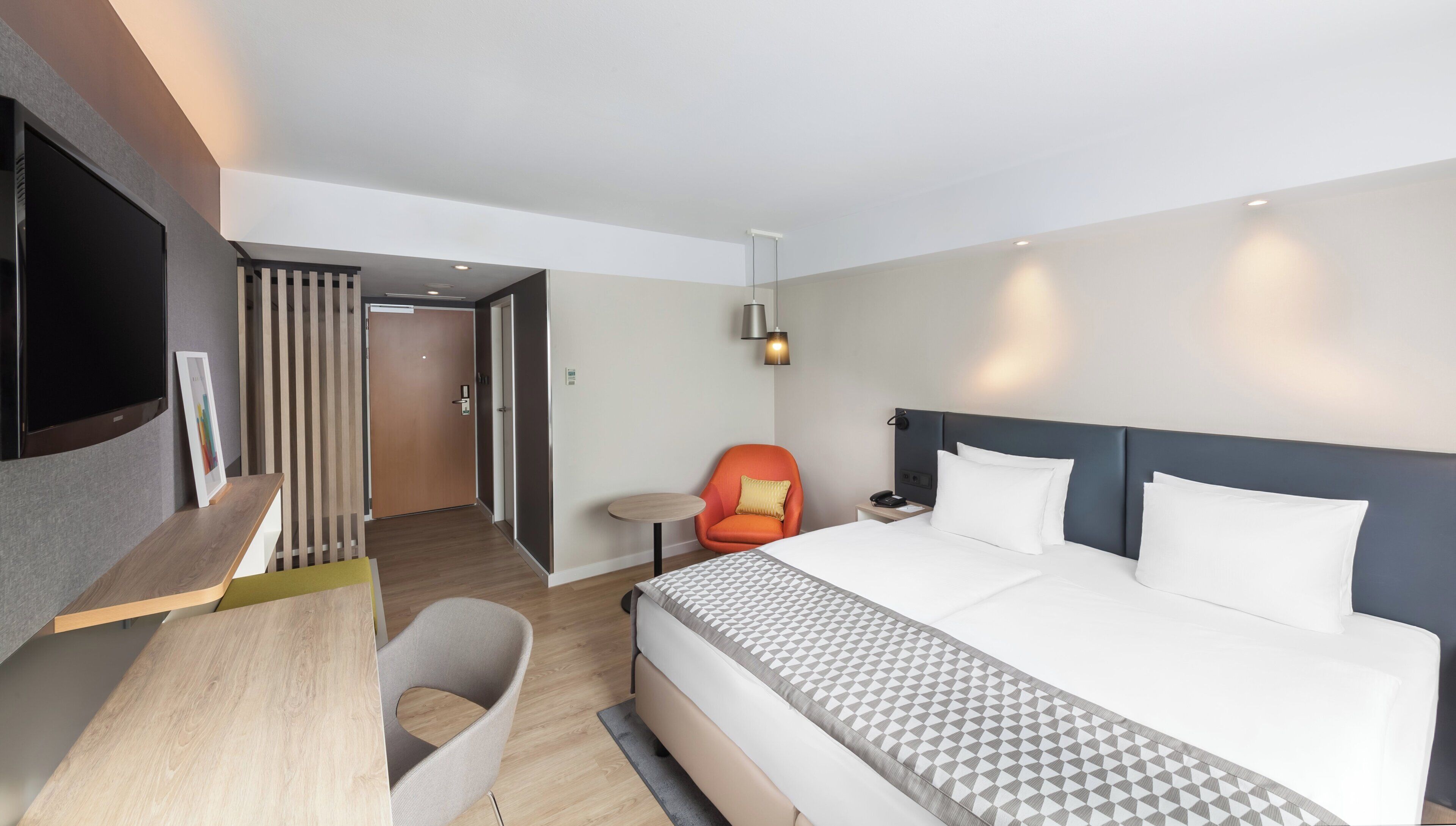 A modern hotel room with a large bed, a desk, a chair, a TV, and a door leading to another room.