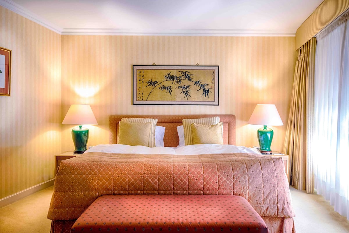 A hotel room with a large bed, two bedside lamps, a framed artwork on the wall, and a window with curtains.