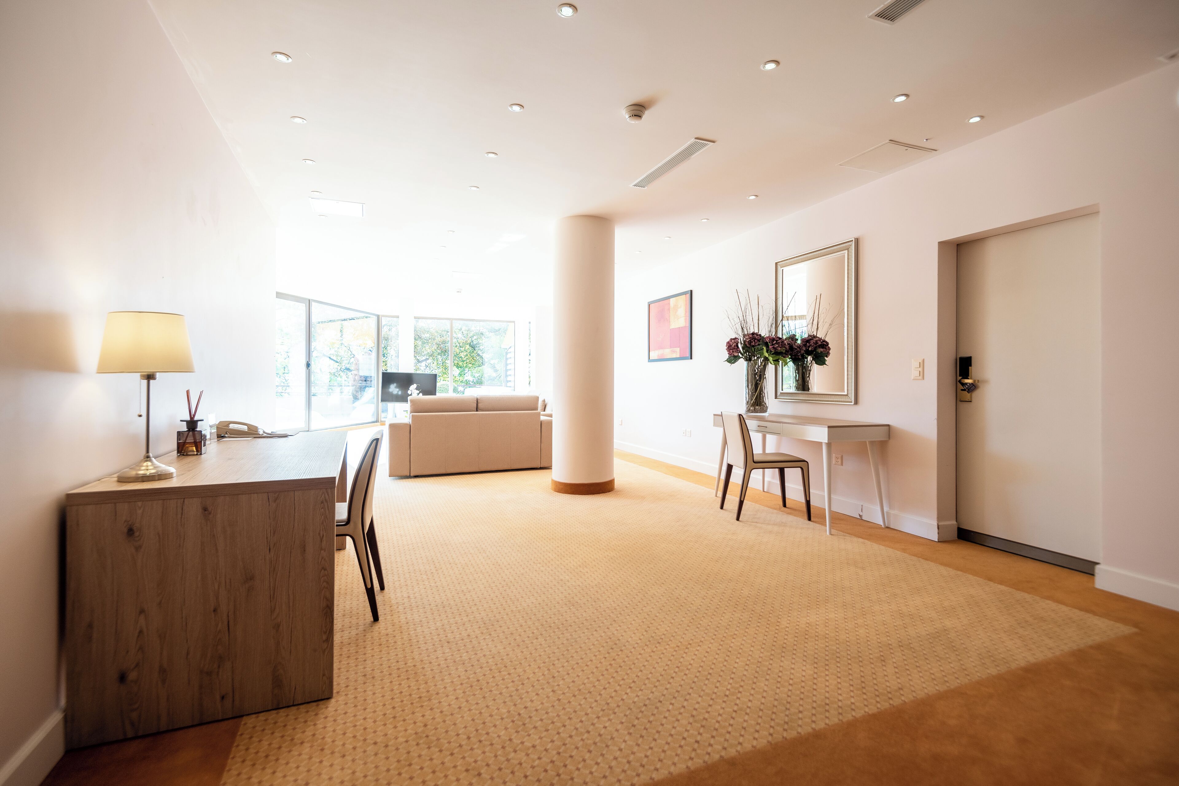 Un hall d’hôtel moderne avec un comptoir d’accueil en bois, un canapé beige, un fauteuil blanc, une petite table blanche ornée d’un vase de fleurs et un grand miroir au mur.