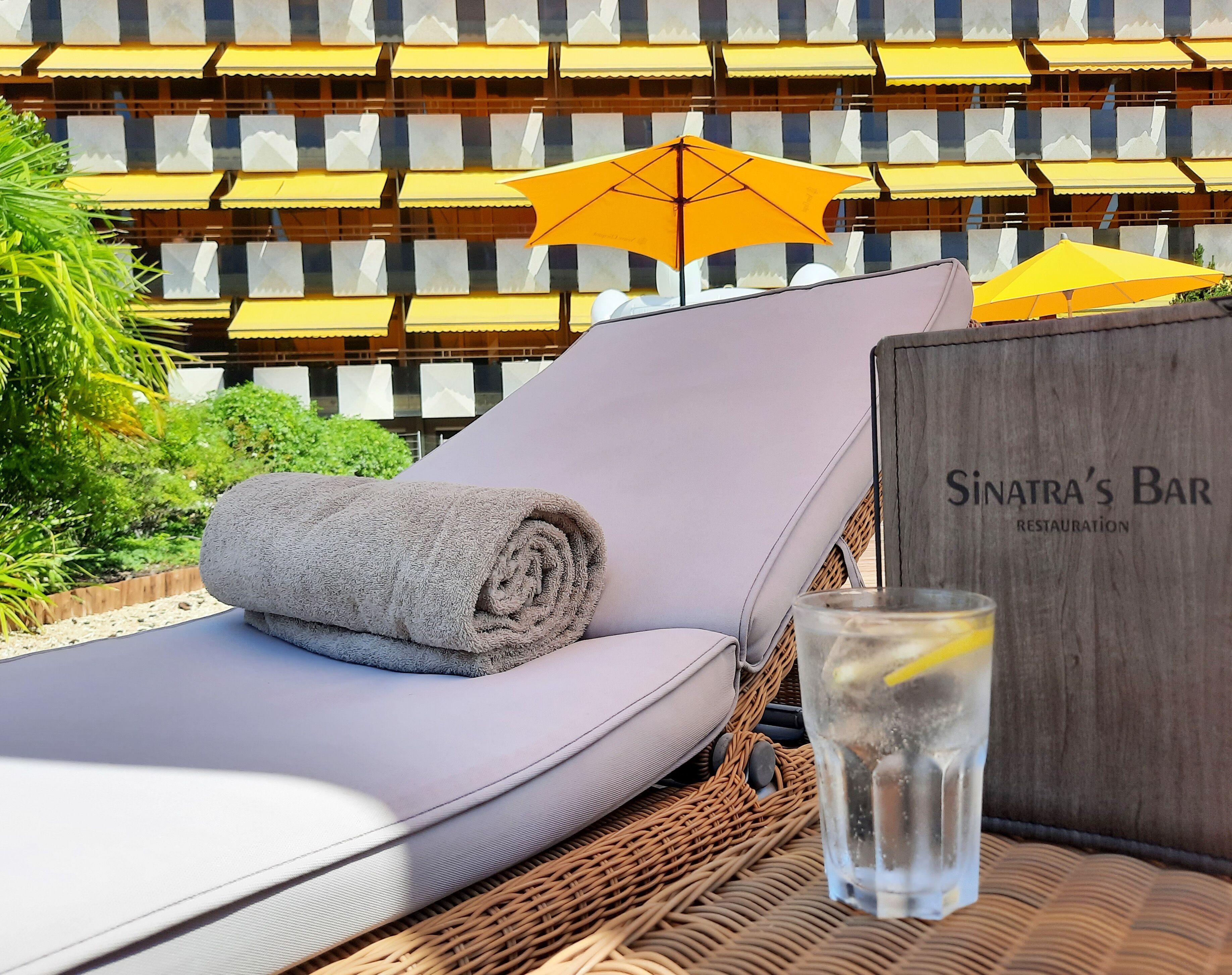 A sun lounger with a rolled towel, a glass of water with a lemon slice, and a wooden sign for Sinatra's Bar.