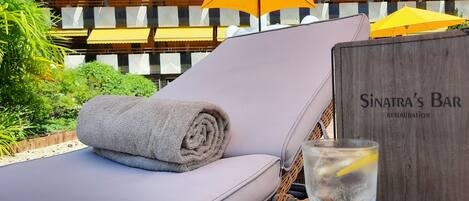 A sun lounger with a rolled towel, a glass of water with a lemon slice, and a wooden sign for Sinatra's Bar.