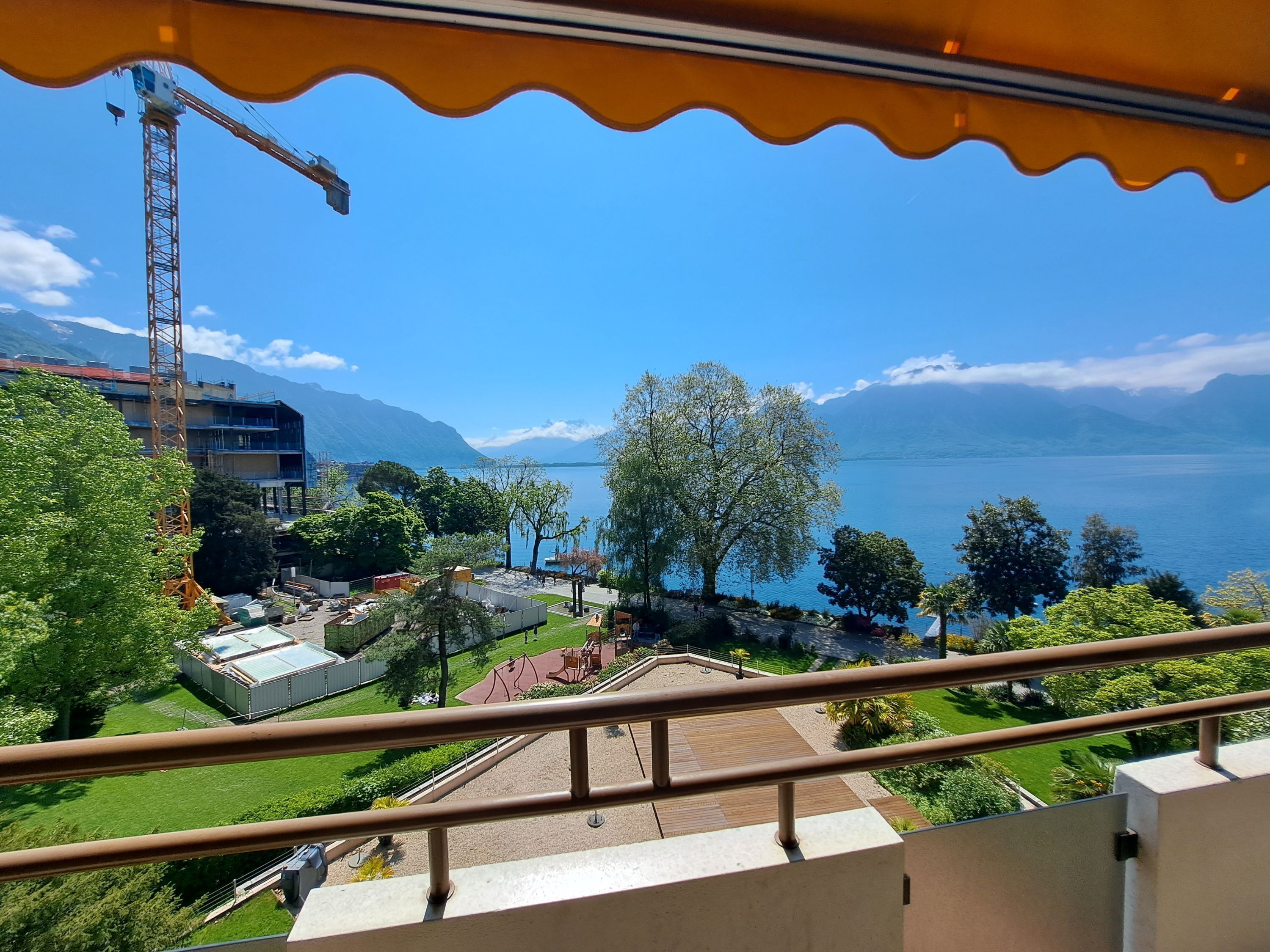 A balcony with a view of a construction crane, a playground, and a lake with mountains in the background.