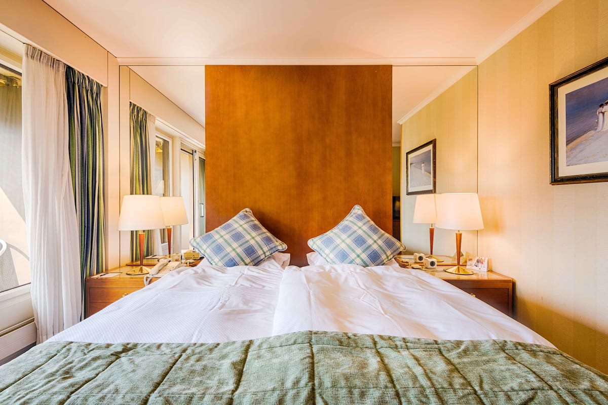 A hotel room with a large bed, two bedside tables with lamps, a wooden headboard, and a window with curtains.