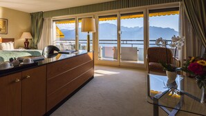 Habitación de hotel con cama, escritorio con lámpara, silla, mesa de vidrio con un jarrón de flores y vistas a las montañas a través de grandes ventanales.