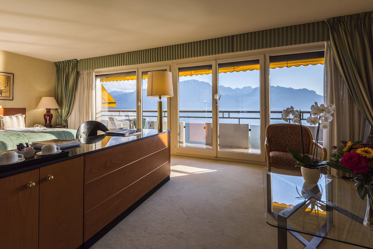 A hotel room with a bed, a desk with a lamp, a chair, a glass table with a vase of flowers, and a view of mountains through large windows.