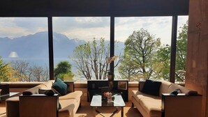 Una sala de estar moderna con un ventanal que ofrece vistas a montañas y una masa de agua. Hay dos sofás, una mesa de centro y una mesita auxiliar con objetos decorativos.