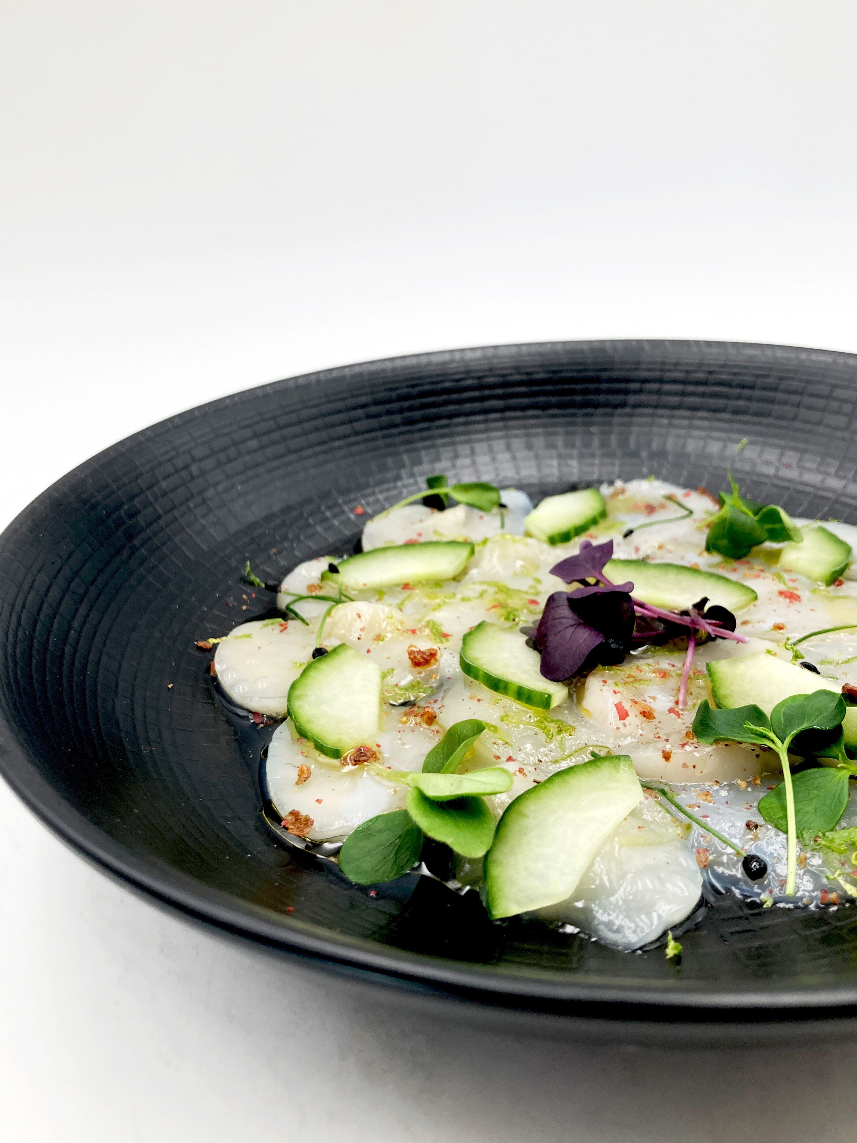 Un plato con rodajas de pepino, verduras de hoja verde y posiblemente pescado, sobre un plato negro.