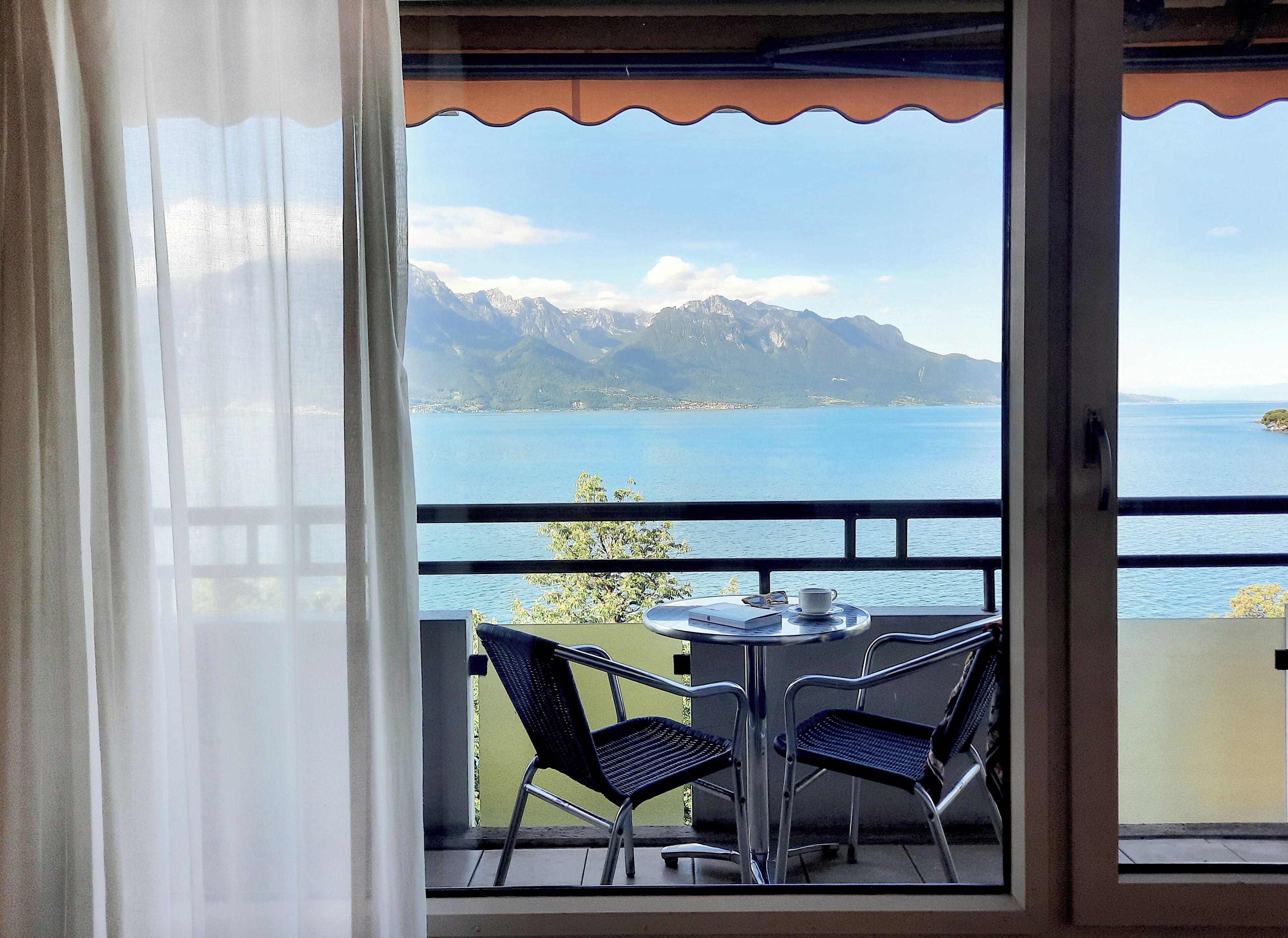 Un balcon avec deux chaises et une table, offrant une vue sur un paysage montagneux et une étendue d’eau.