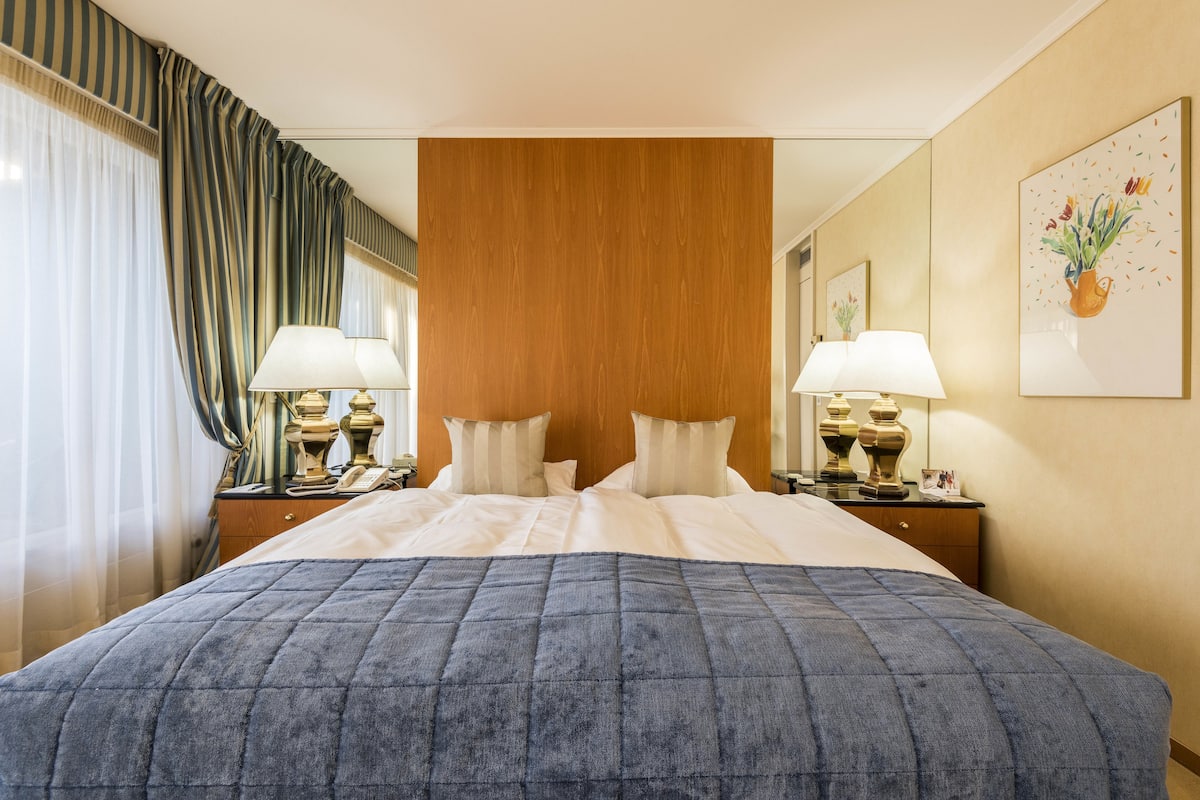 A hotel room with a large bed, two bedside lamps, a wooden headboard, a painting on the wall, and a window with curtains.