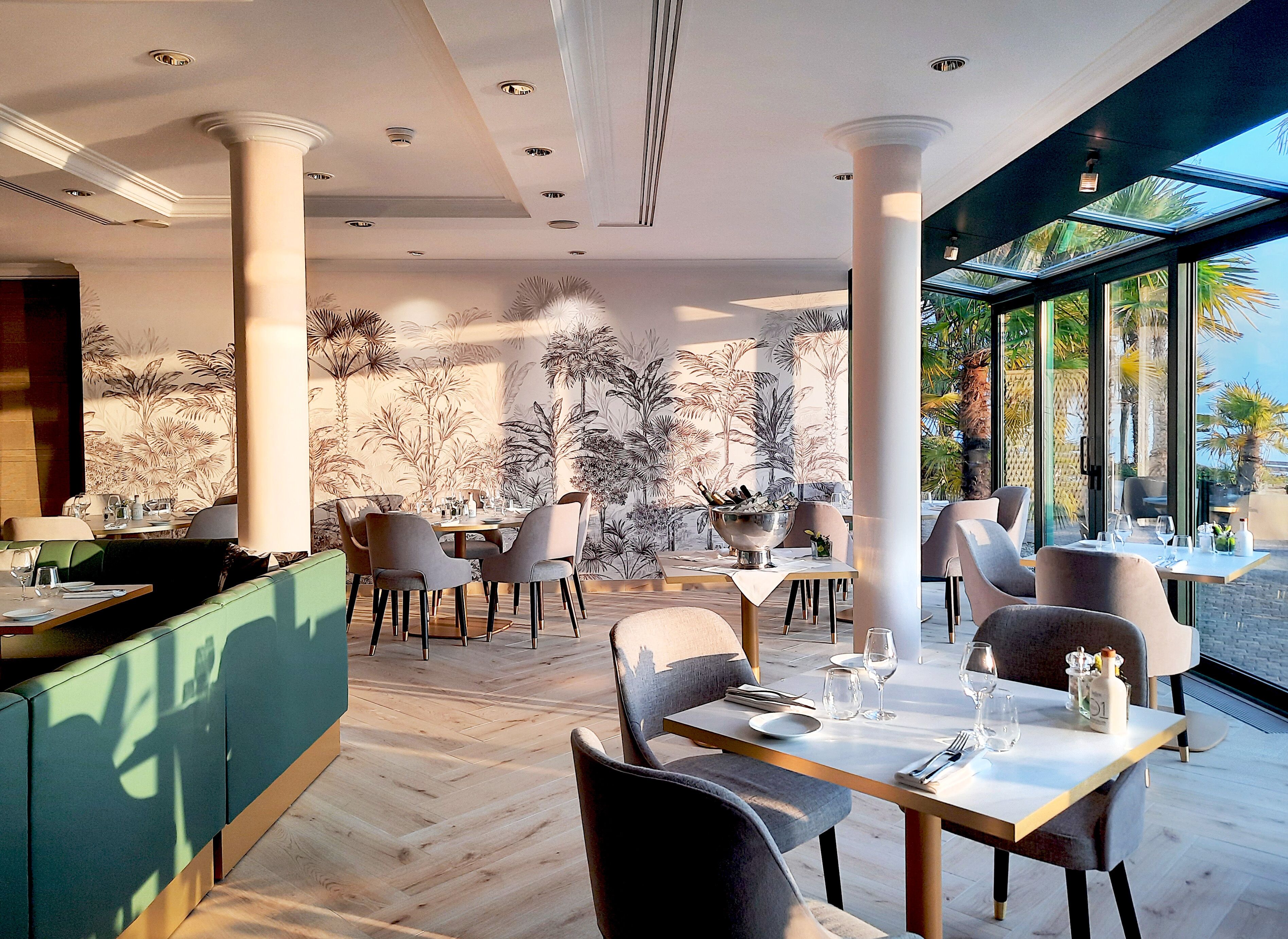 Un espace de restauration moderne, doté d’un plafond haut, de grandes fenêtres et d’une fresque murale aux motifs tropicaux.