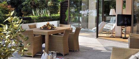 A modern outdoor dining area with wicker furniture, a glass-enclosed TV, and lush greenery.