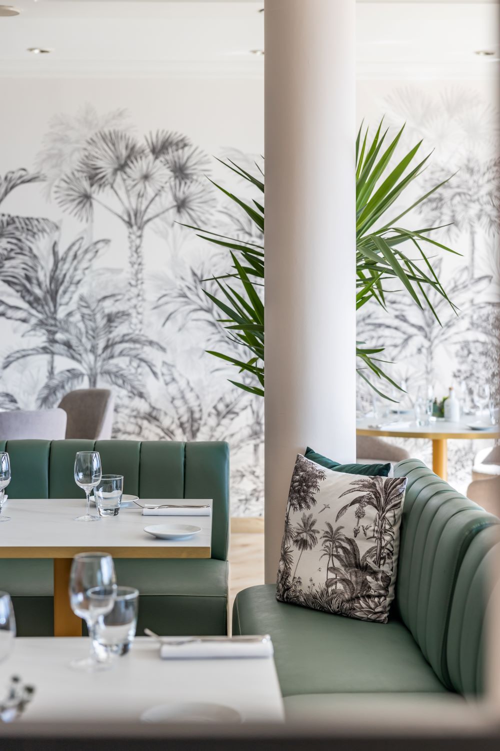 Un área de comedor con un sofá verde, una mesa con vasos y platos, y una pared con un mural de plantas tropicales.