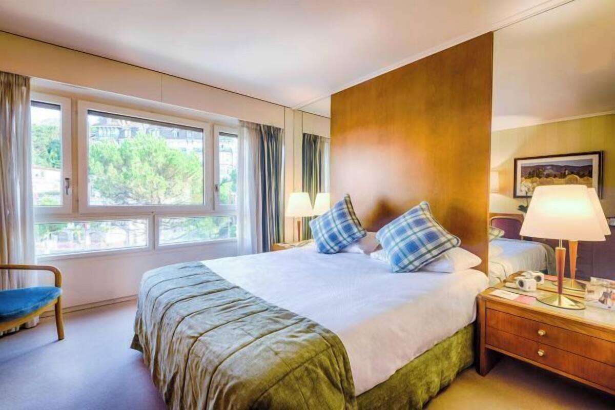 Una habitación de hotel con una cama, dos lámparas de noche, una mesita de noche, una silla y una ventana con cortinas.