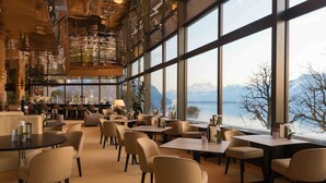 Amplia zona de comedor con grandes ventanales que ofrecen vistas a montañas y un lago. Se disponen mesas y sillas para comer.