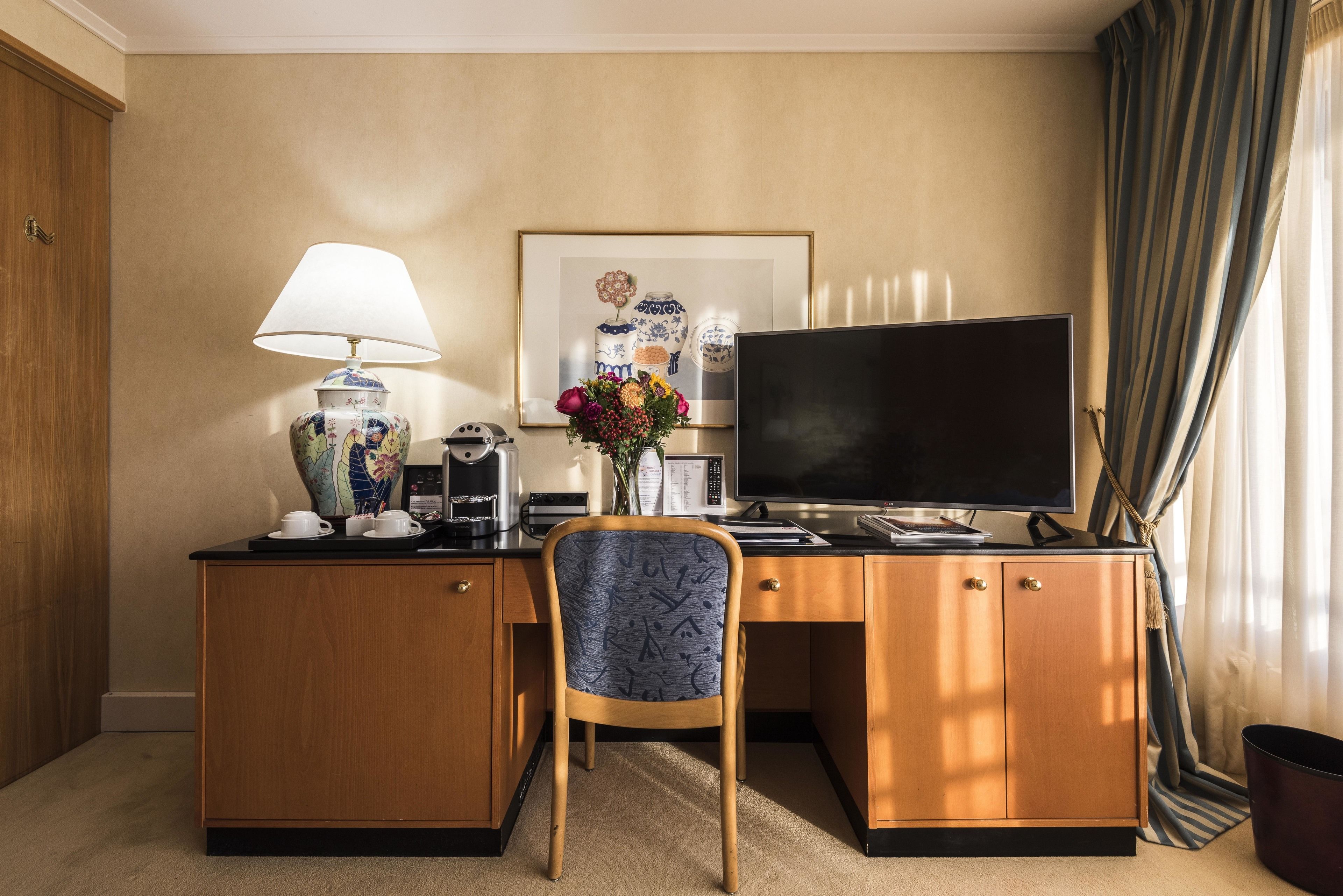 Habitación de hotel con escritorio de madera, televisor, lámpara, un florero con flores y un cuadro en la pared.