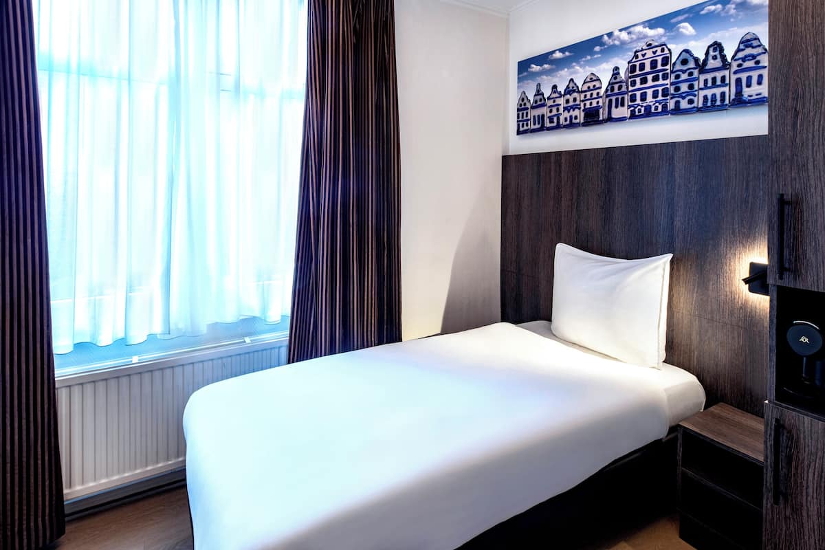 Una habitación de hotel con cama, ventana con cortinas, un cuadro en la pared que muestra edificios y cabecero de madera.