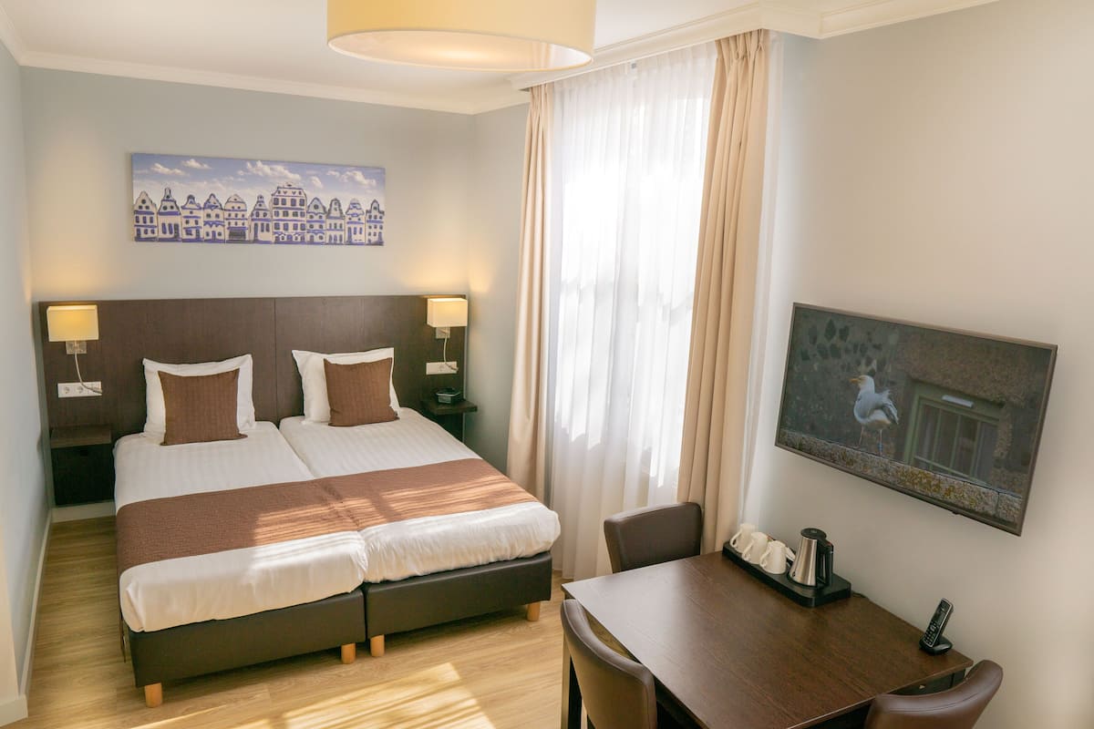 Habitación de hotel con cama doble, mesitas de noche con lámparas, un escritorio con silla, un cuadro en la pared y una ventana con cortinas.