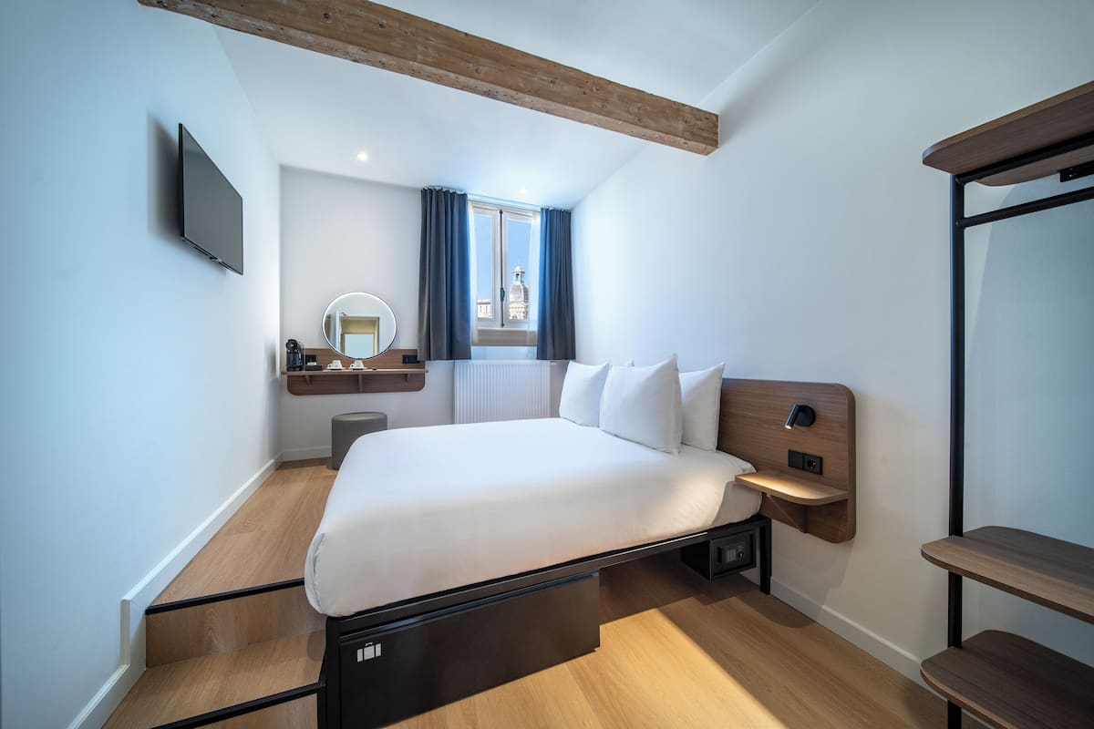 Un dormitorio moderno con cama, mesitas de noche, televisor, ventana con cortinas y vista a un edificio.