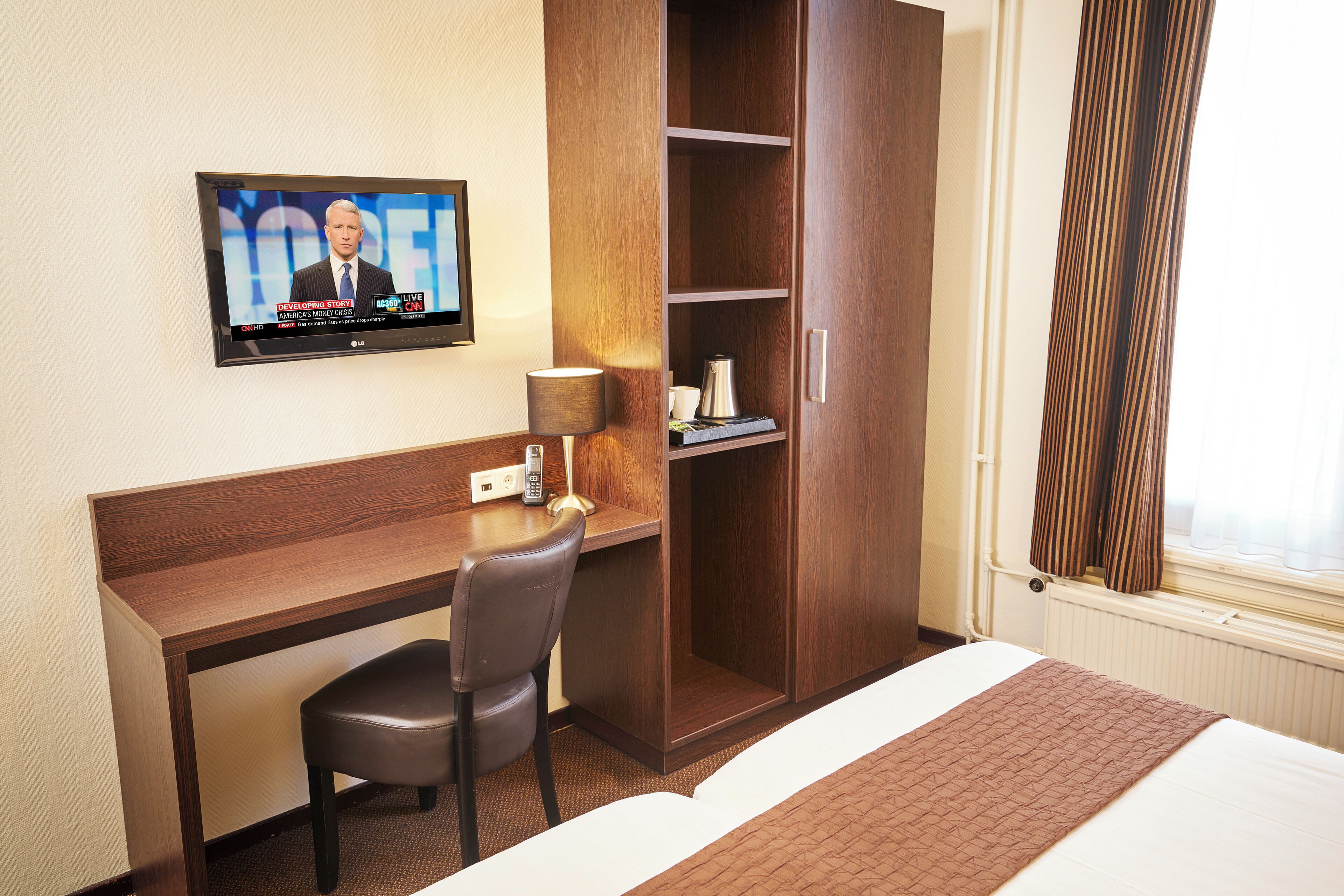 Quarto de hotel com escrivaninha, cadeira, televisão, guarda-roupa e uma cama.