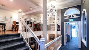 A grand staircase with white balustrades leading to a second floor, a dining area with wooden chairs and tables, and a hallway with a blue carpet and decorative ceiling lights. - Hotel Nes Amsterdam (Amsterdam)
