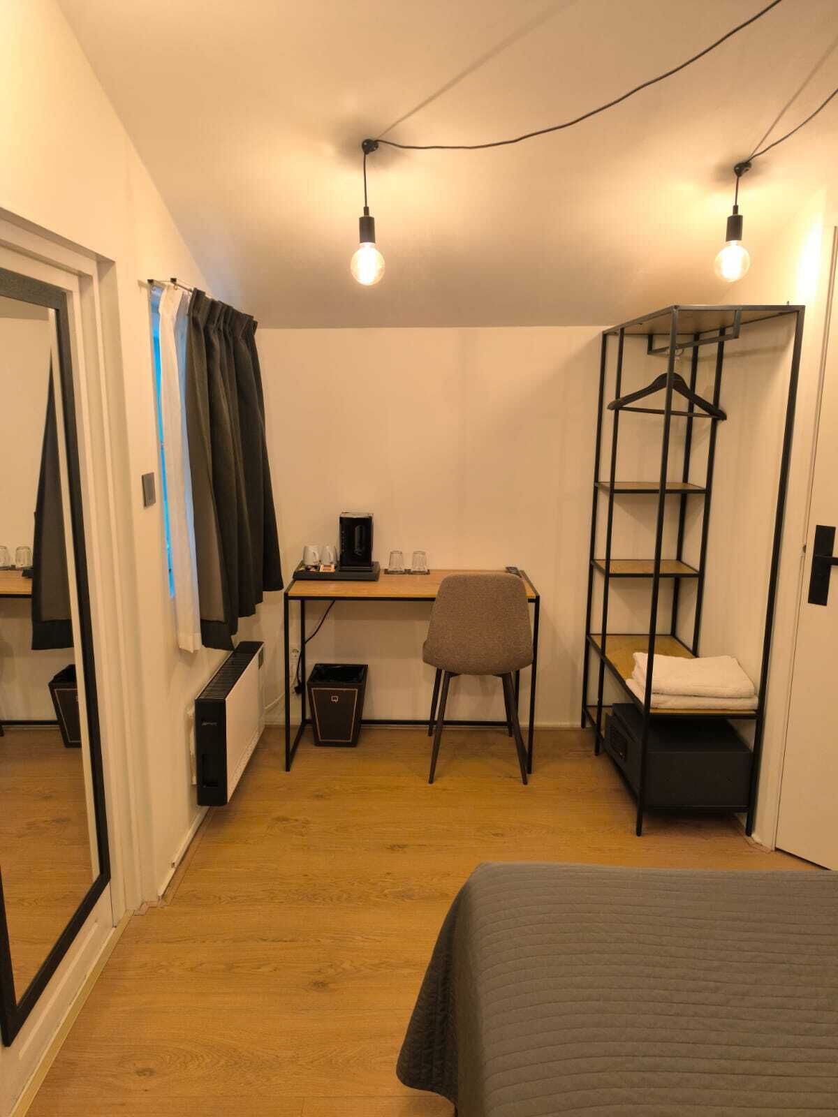 A compact room with a bed, a desk, a chair, a radiator, a hanging clothes rack, and a wardrobe.
