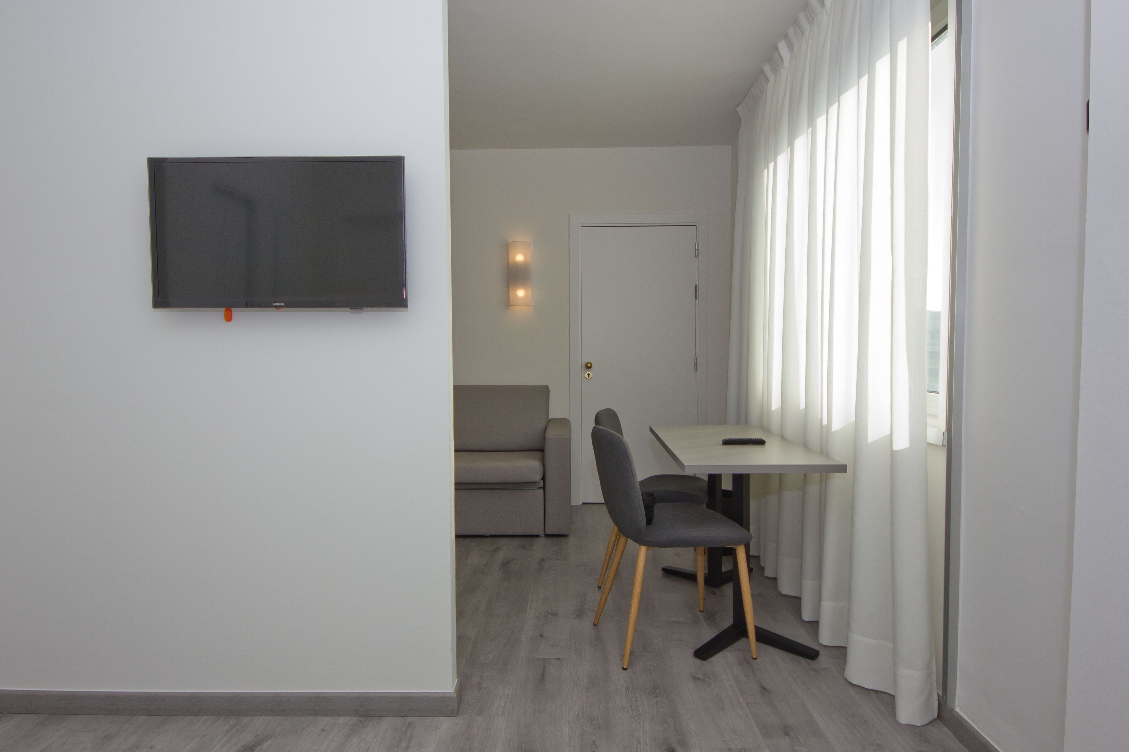 Ein modernes Wohnzimmer mit einem an der Wand befestigten Flachbildfernseher, einem grauen Sofa, einem Esstisch mit Stühlen und einem großen Fenster mit weißen Vorhängen.