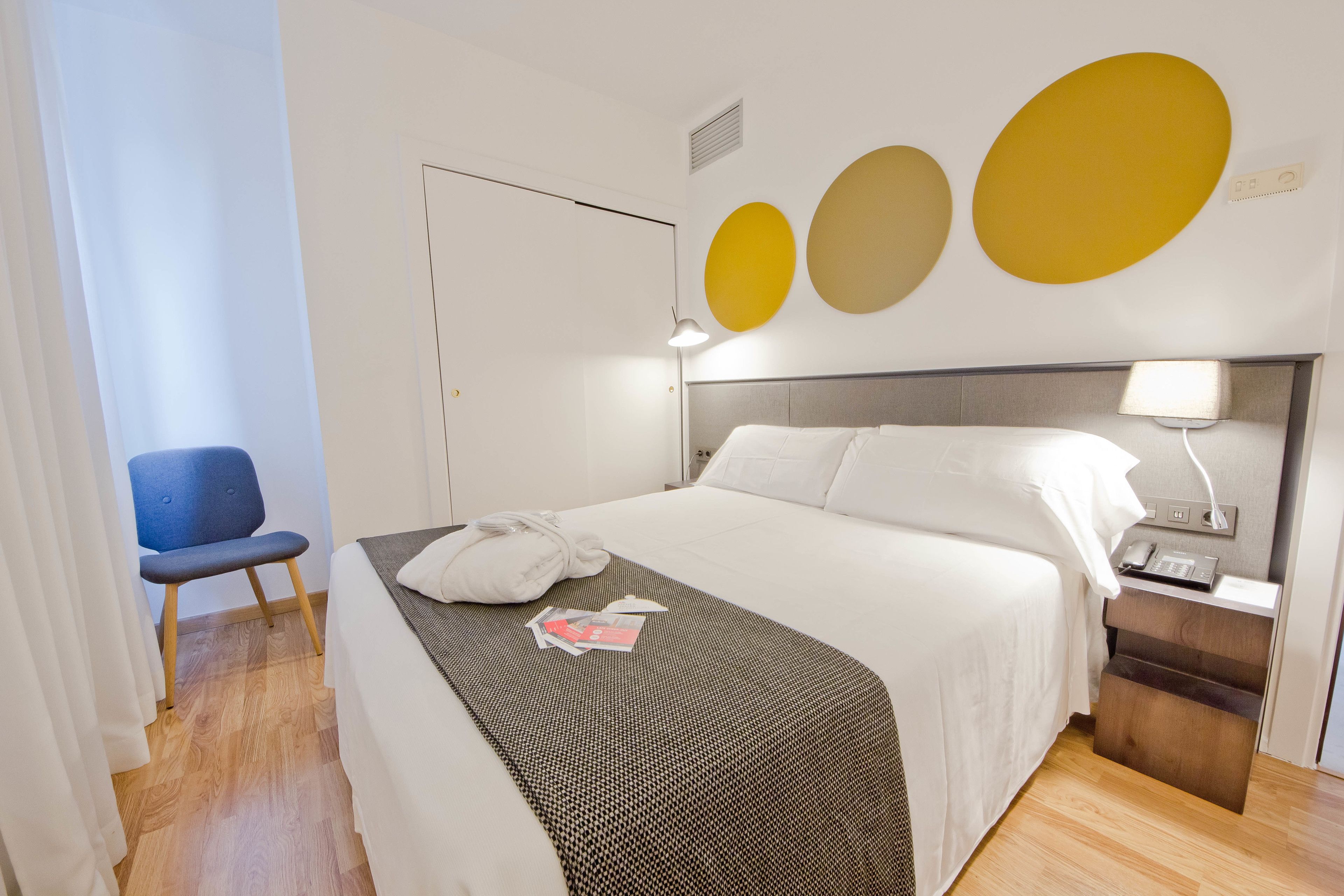 Ein ordentlich eingerichtetes Hotelzimmer mit Bett, Nachttisch, Lampe und einem Sessel.