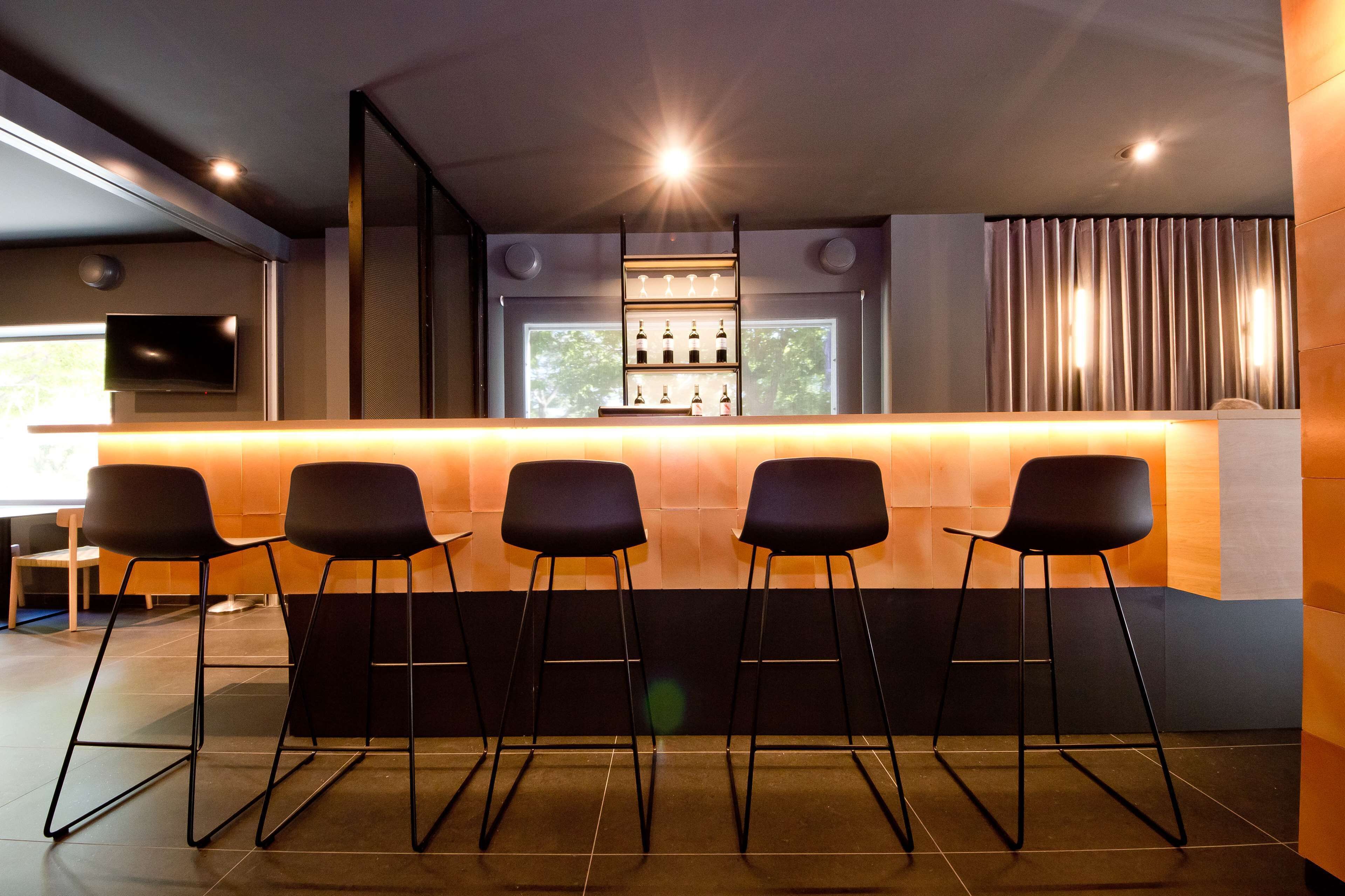 Una zona de bar con cuatro taburetes negros, una estantería con botellas y un televisor en la pared.